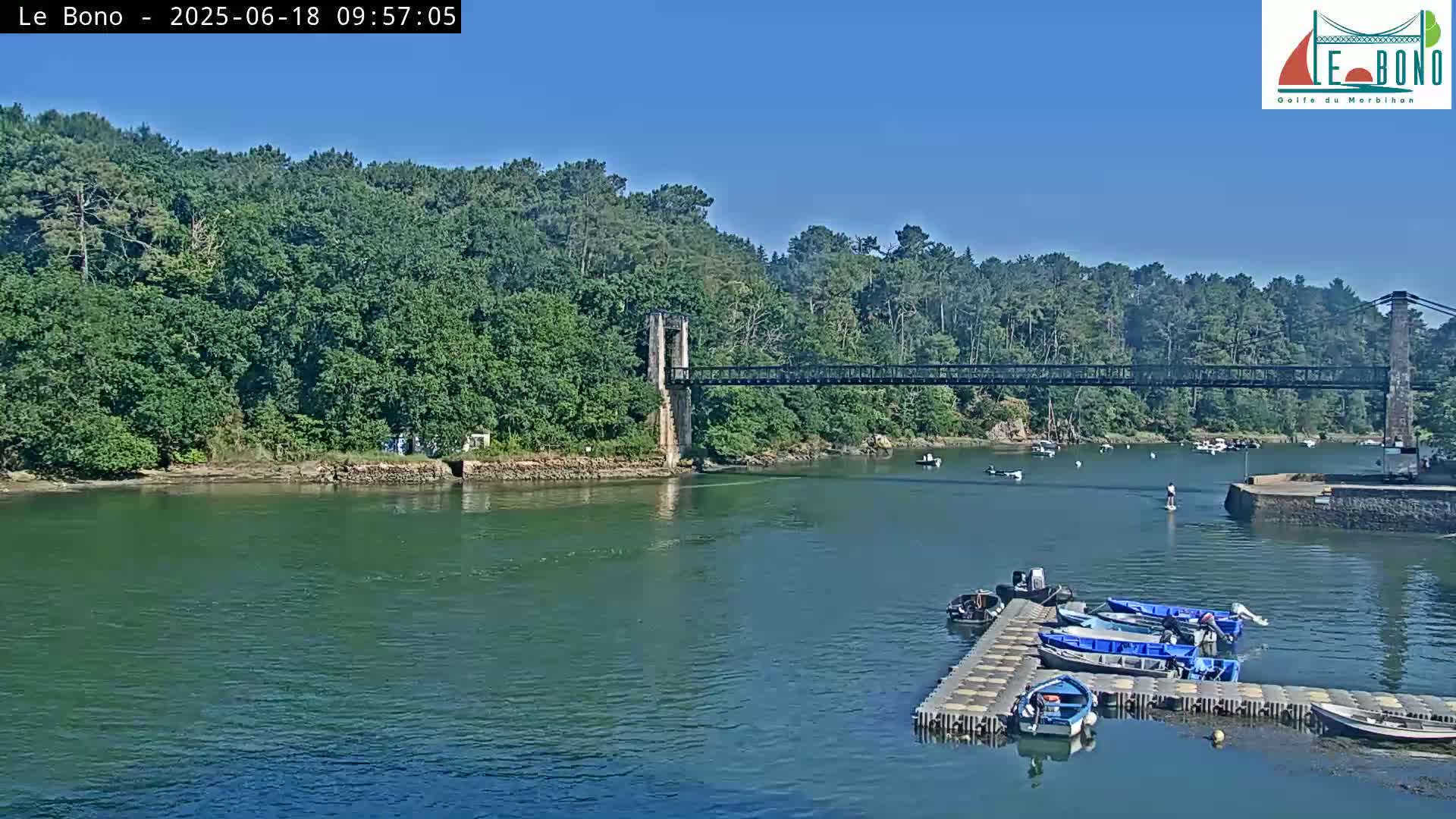 A suspension bridge spans a calm river under a clear blue sky, with several small boats moored at a dock and a few more scattered on the water.