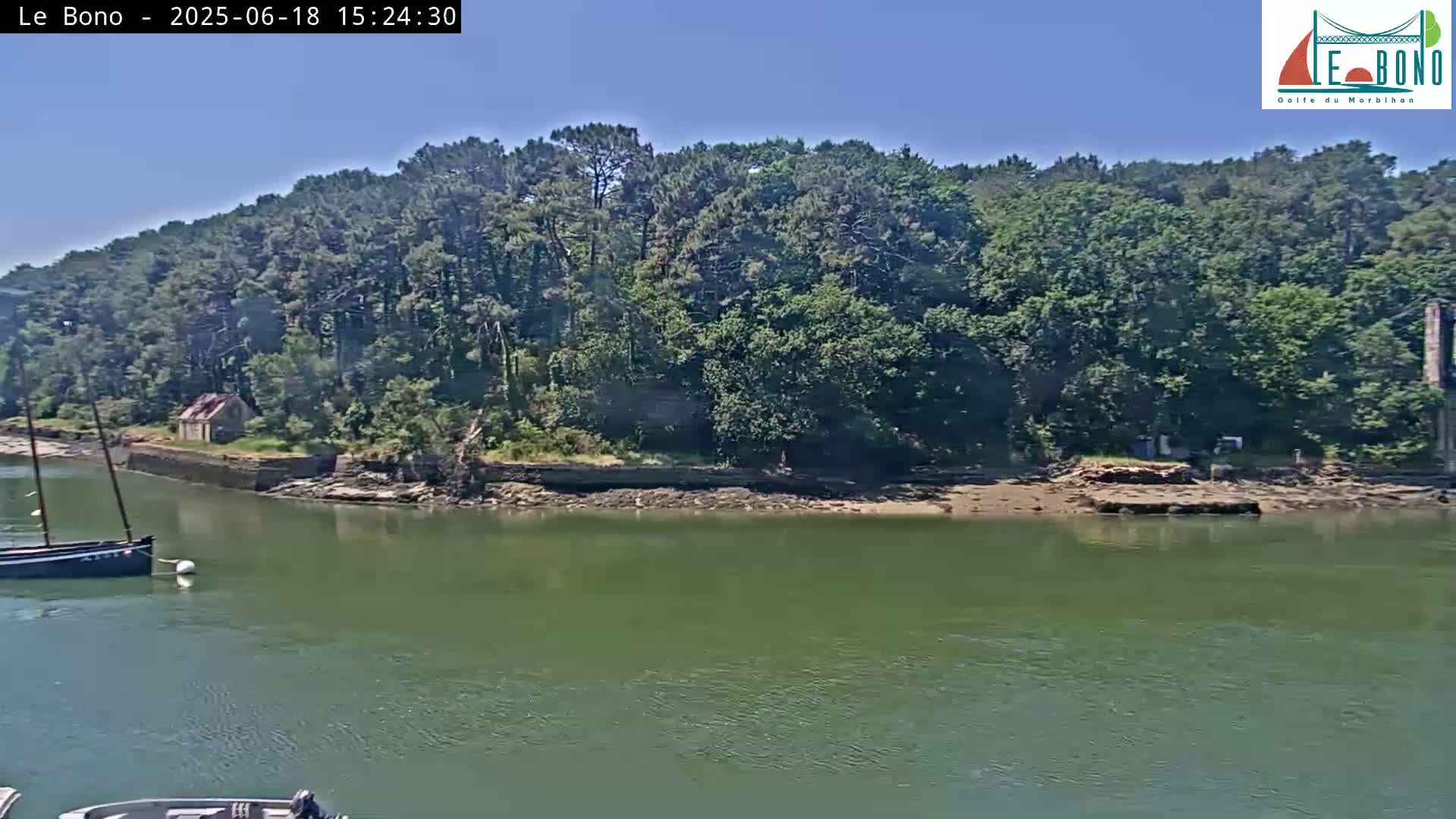 A sailboat is moored near a rocky shoreline lined with lush green trees under a clear blue sky.