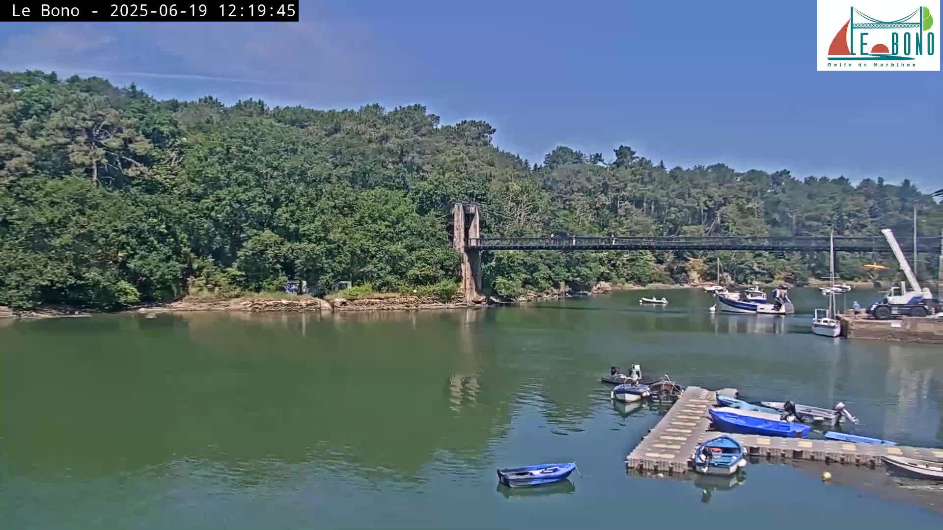 A suspension bridge spans a calm river, lined with green trees and several small boats moored at a dock under a clear blue sky.