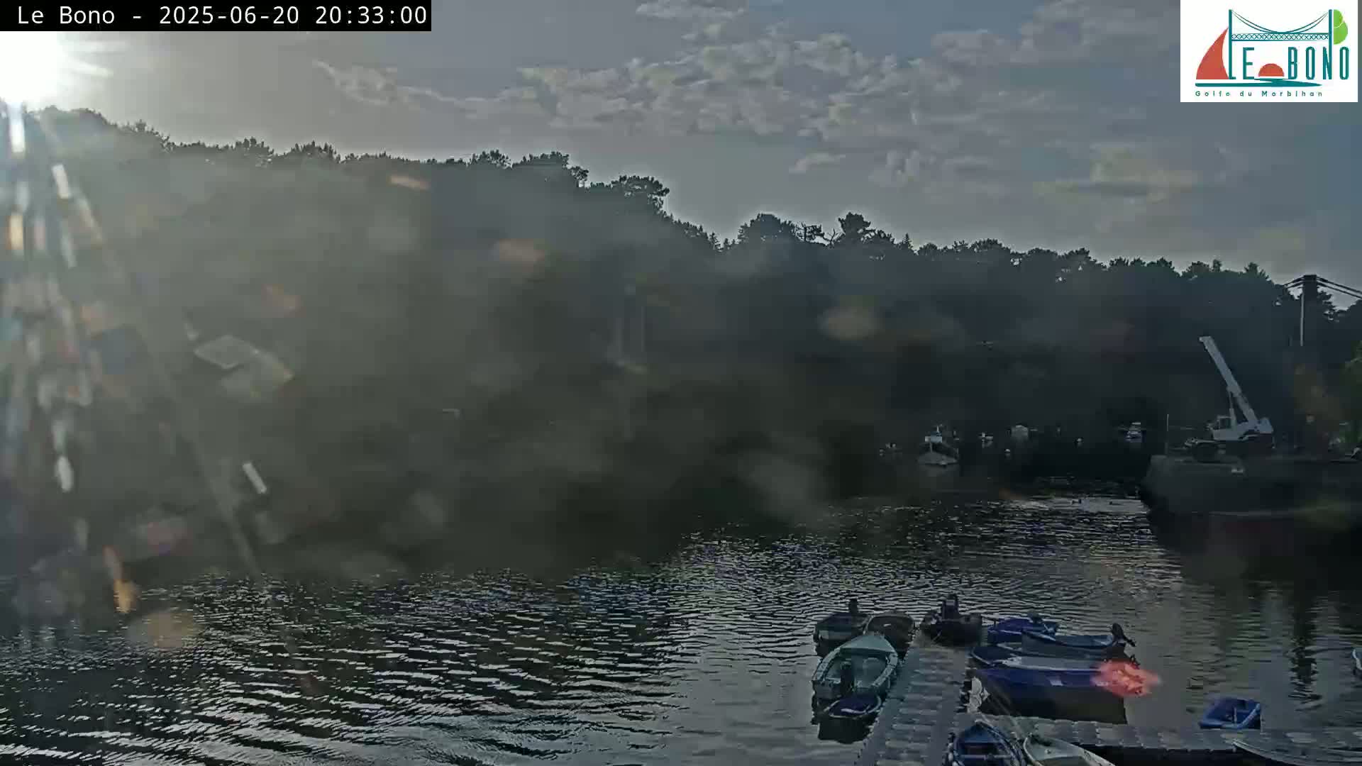 Several small boats are moored at a dock near a wooded shoreline under a partly cloudy sky.