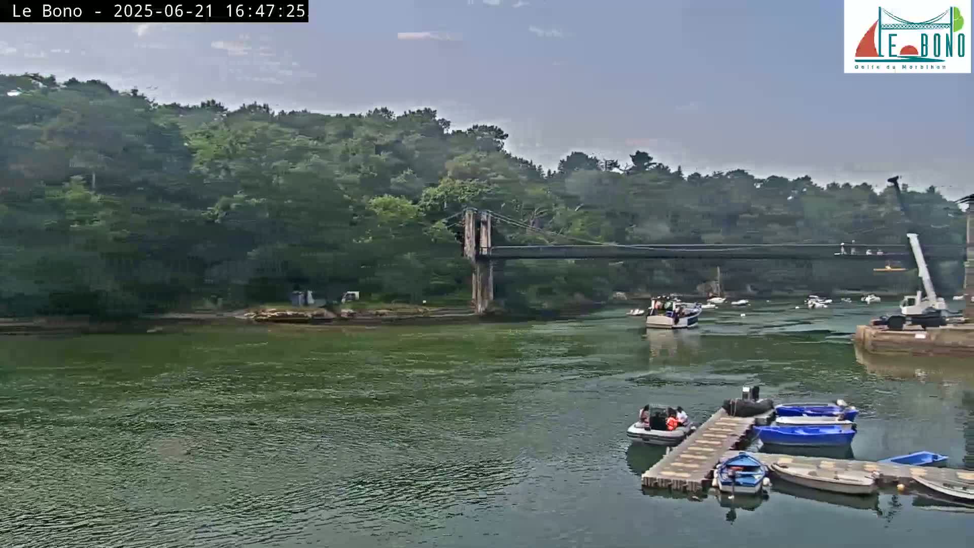 A suspension bridge spans a calm, green river, with several small boats moored at a dock beneath a mostly clear sky.