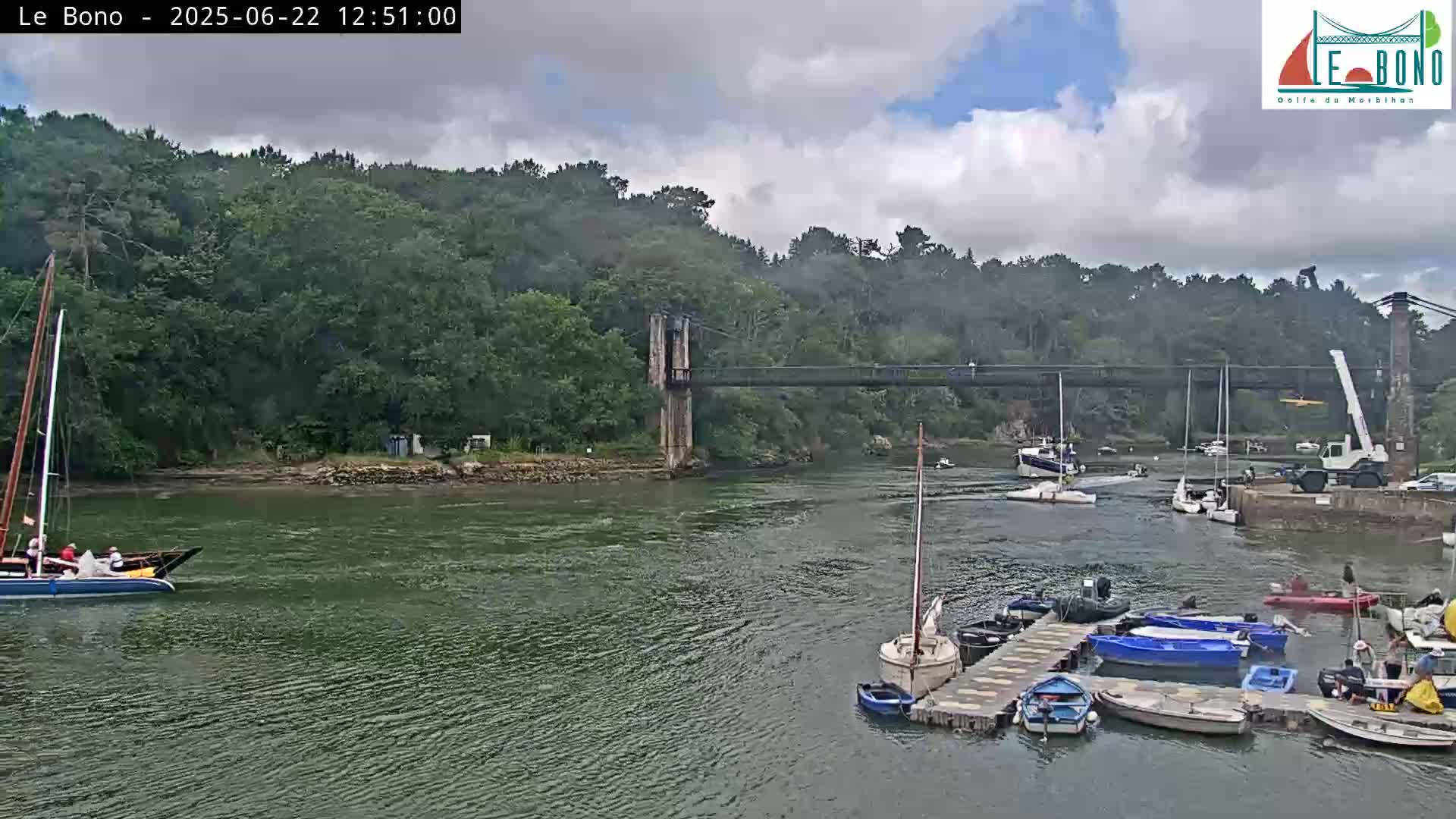 A suspension bridge spans a river with overcast skies, several sailboats and motorboats are on the water near a dock, and a crane is visible on the shore.