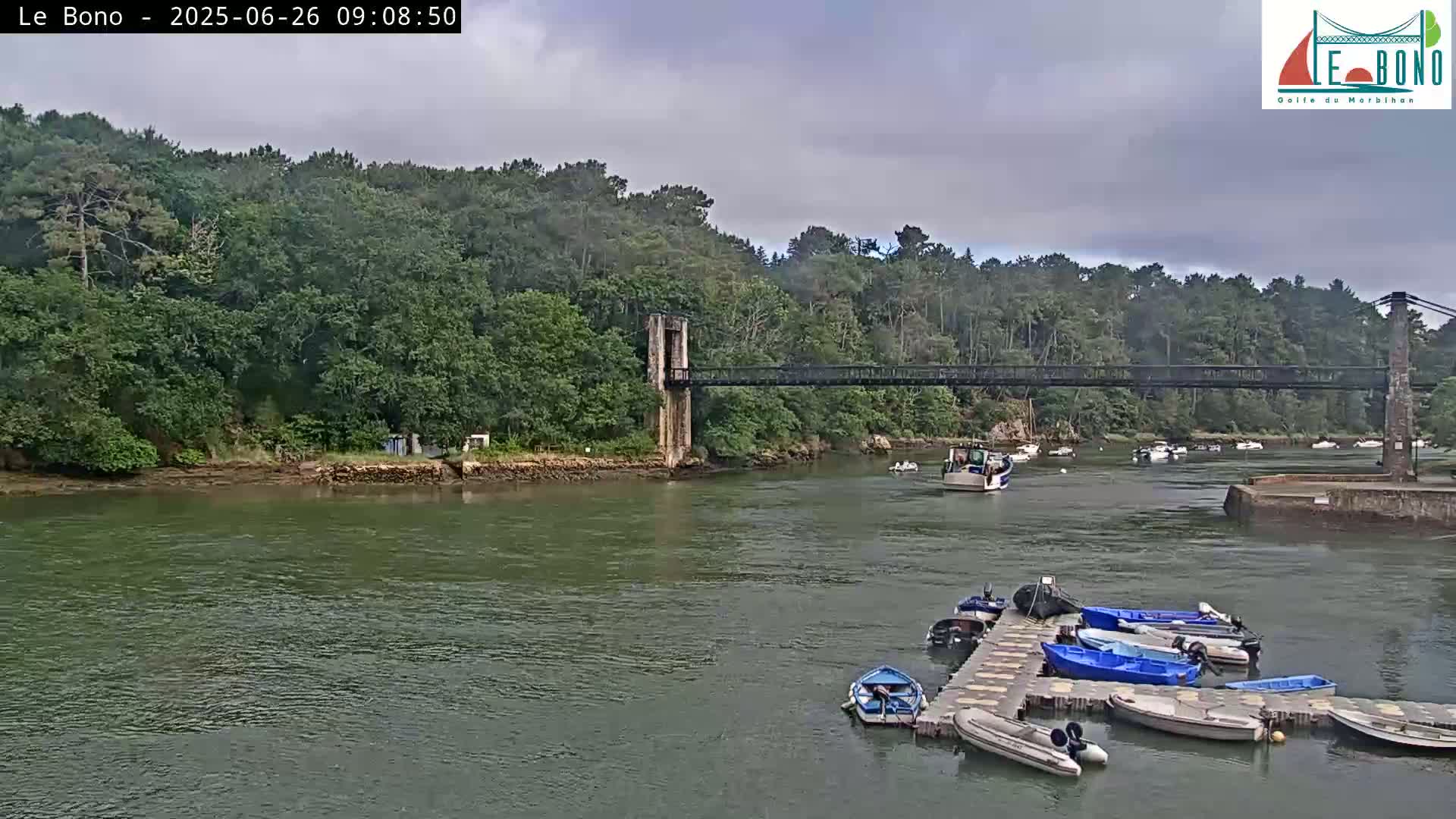 A suspension bridge spans a calm river lined with green trees under an overcast sky, with several boats moored at a dock below.