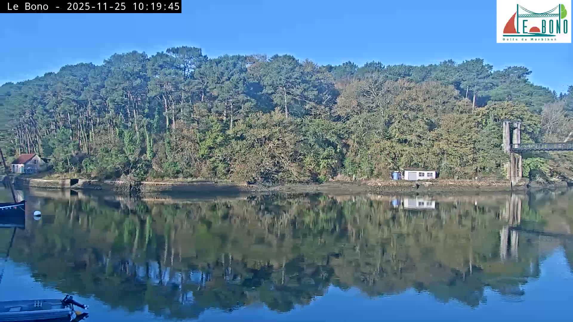 A tranquil river or estuary reflects its densely forested banks, small waterside buildings, and part of a suspension bridge under a clear, sunny sky, with boats partially visible in the foreground.