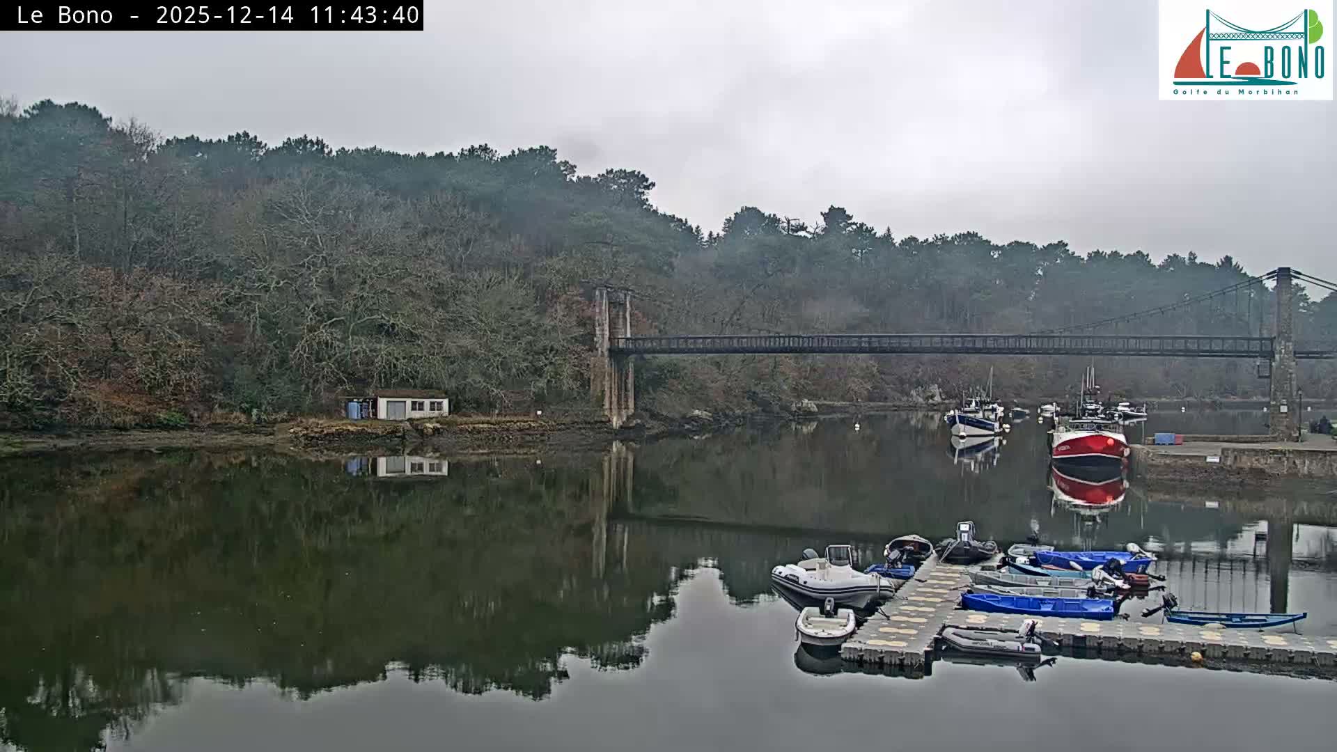 A tranquil river or estuary reflects its densely forested banks, small waterside buildings, and part of a suspension bridge under a clear, sunny sky, with boats partially visible in the foreground.