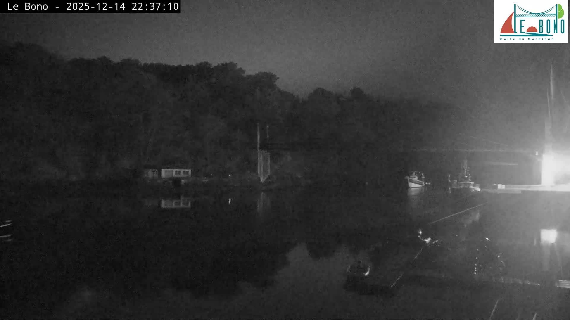 A tranquil river or estuary reflects its densely forested banks, small waterside buildings, and part of a suspension bridge under a clear, sunny sky, with boats partially visible in the foreground.