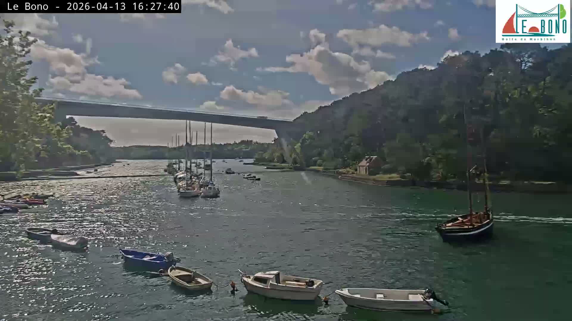A tranquil river or estuary reflects its densely forested banks, small waterside buildings, and part of a suspension bridge under a clear, sunny sky, with boats partially visible in the foreground.
