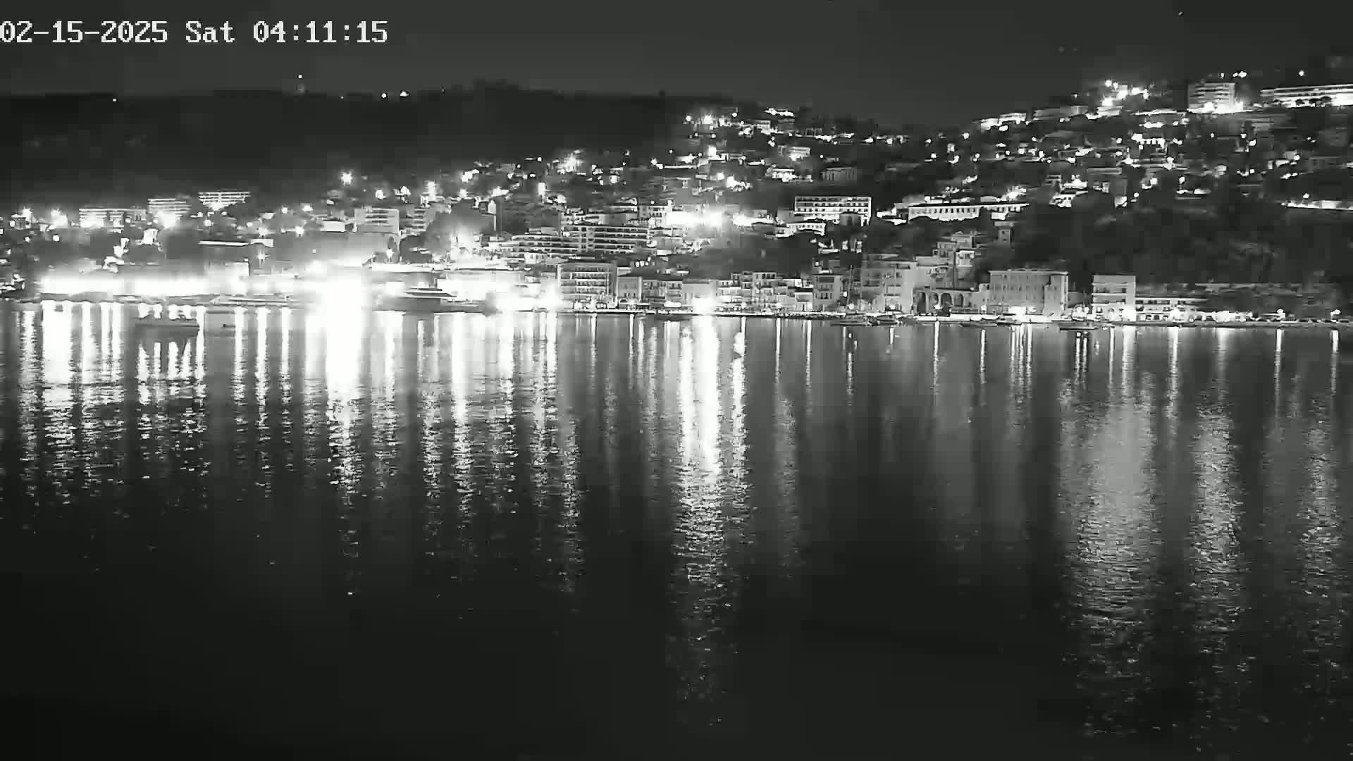 A nighttime, black and white view of a city's waterfront at night, with the lights of buildings and boats reflected in calm water.