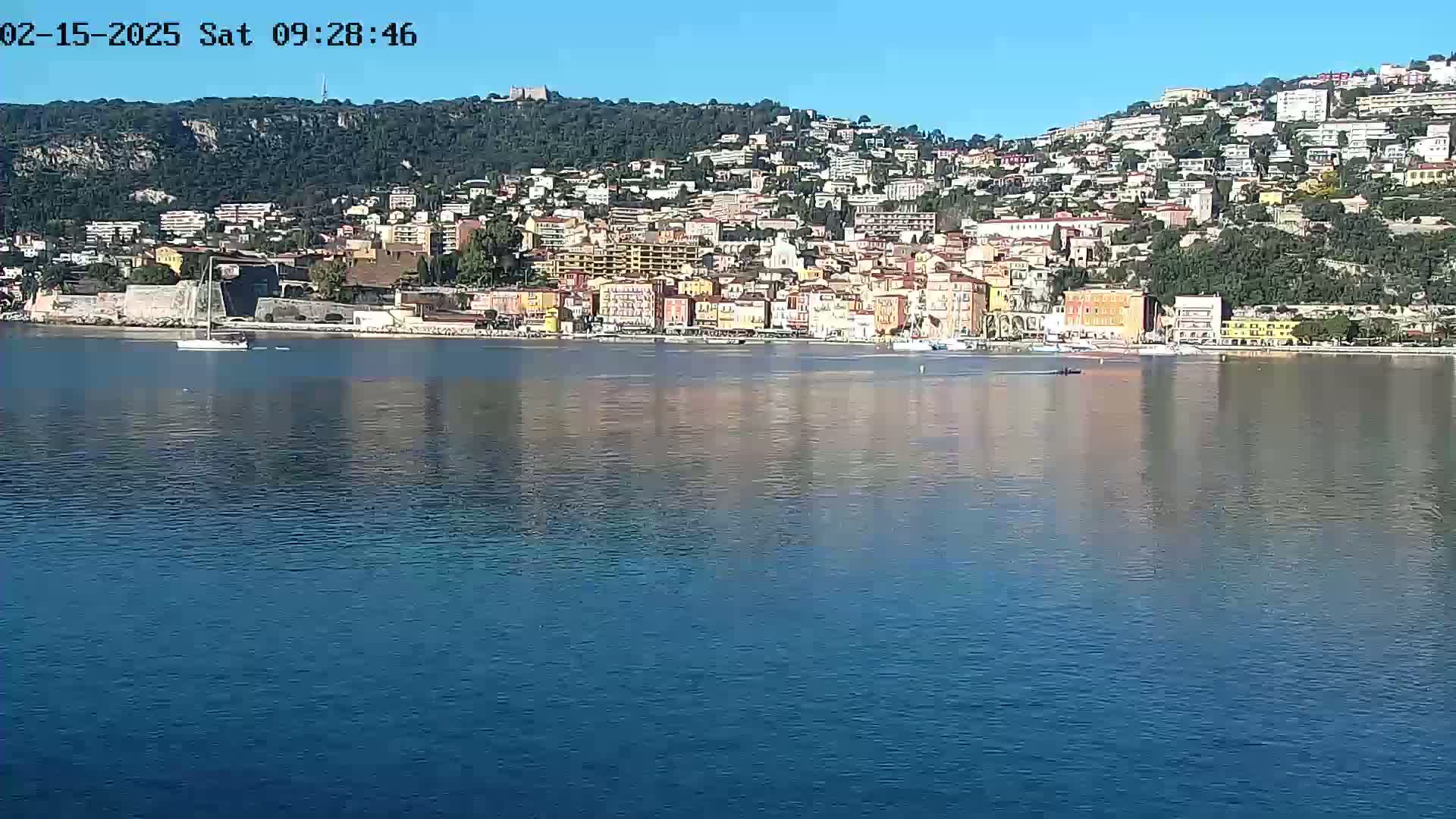 A colorful coastal town sits on a hillside overlooking calm, blue water under a clear sky.