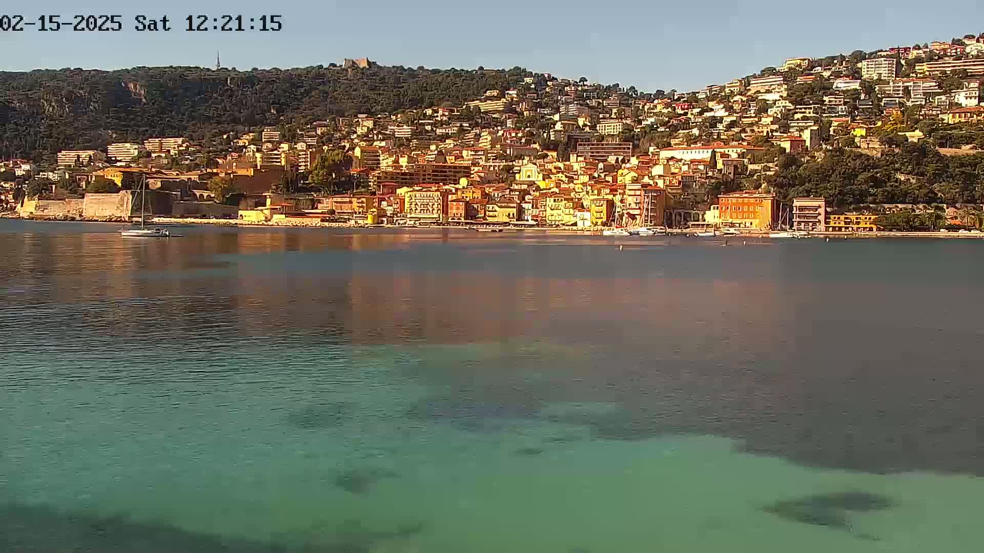 A calm, clear day reveals a coastal town nestled on a hillside, its colorful buildings stretching along the waterfront, with a sailboat visible in the bay.