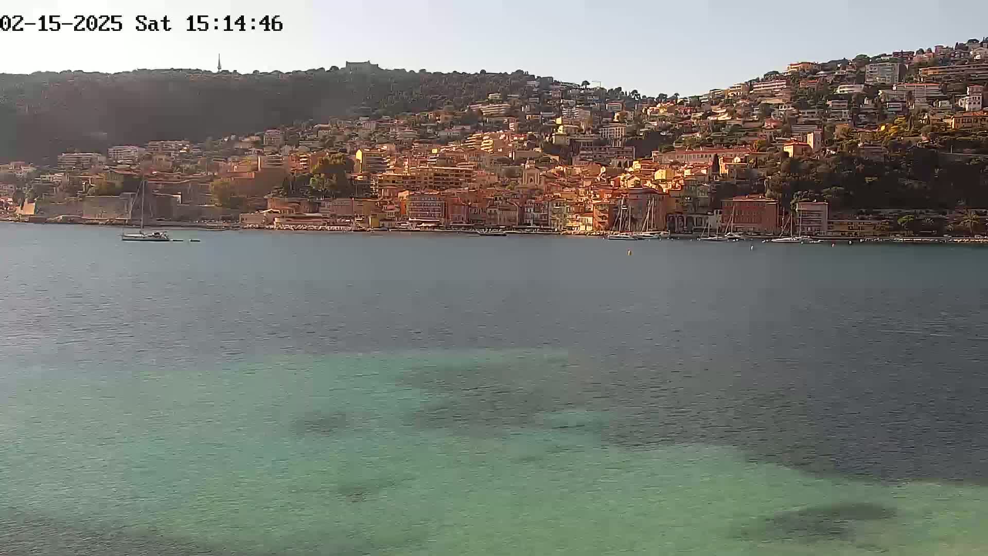 A coastal town with many buildings is situated on a hillside overlooking a calm body of clear, teal water under a mostly clear sky.