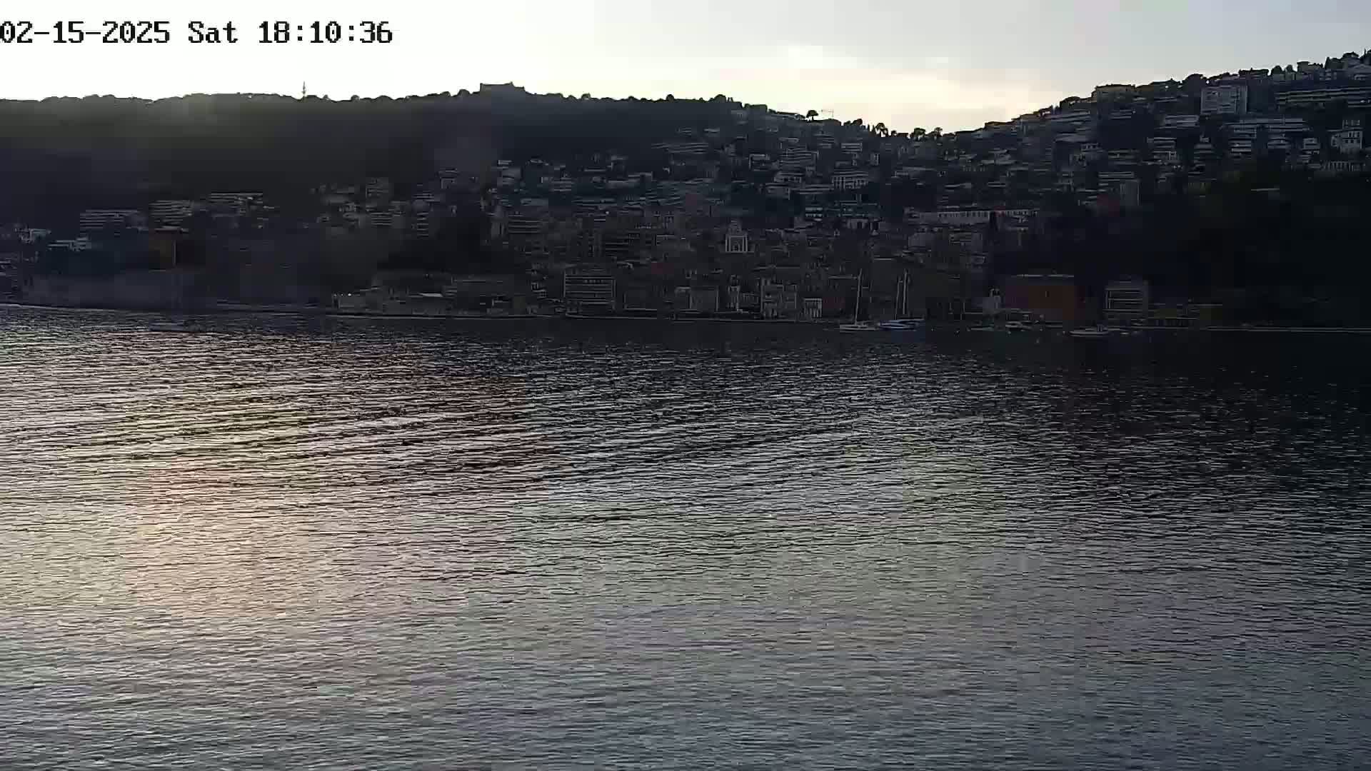 A calm body of water reflects the dusky light of a setting sun, with a hillside town silhouetted against the sky in the background.