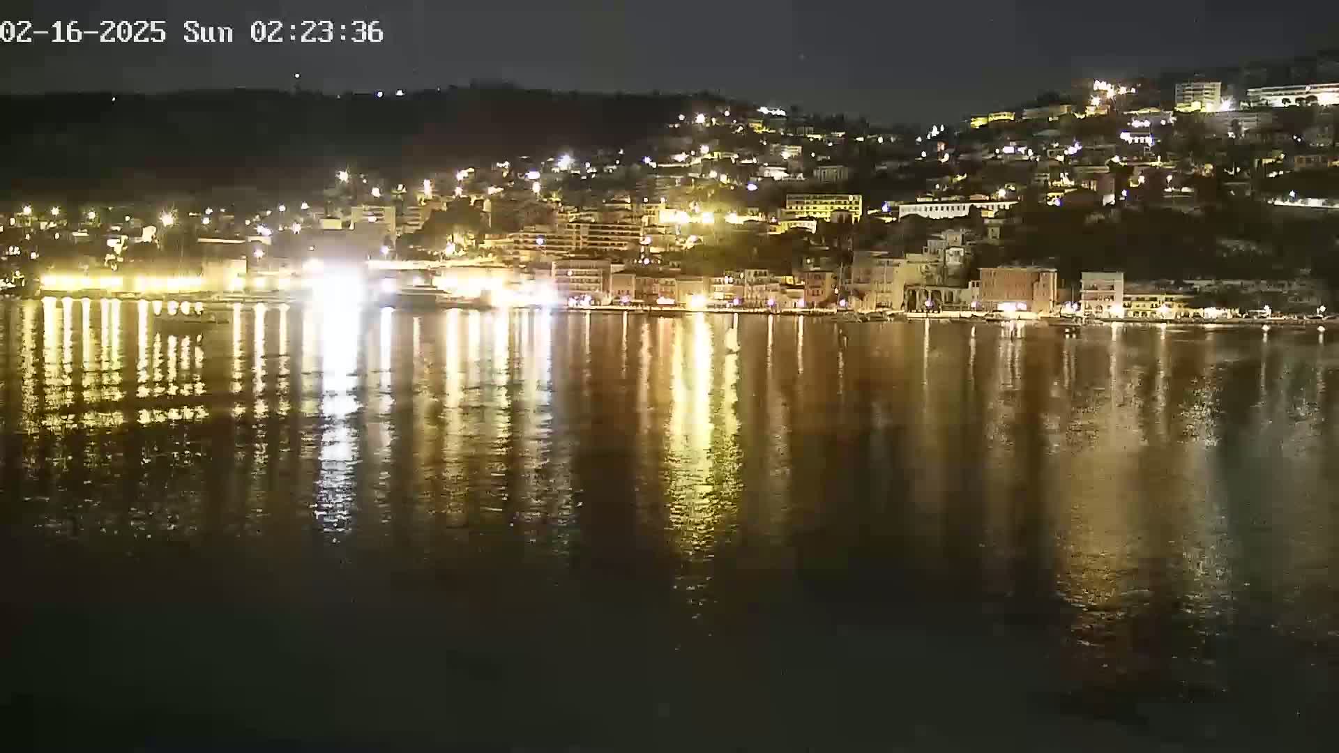 A nighttime view of a waterfront town, brightly lit buildings reflected in a calm body of water under a clear sky.