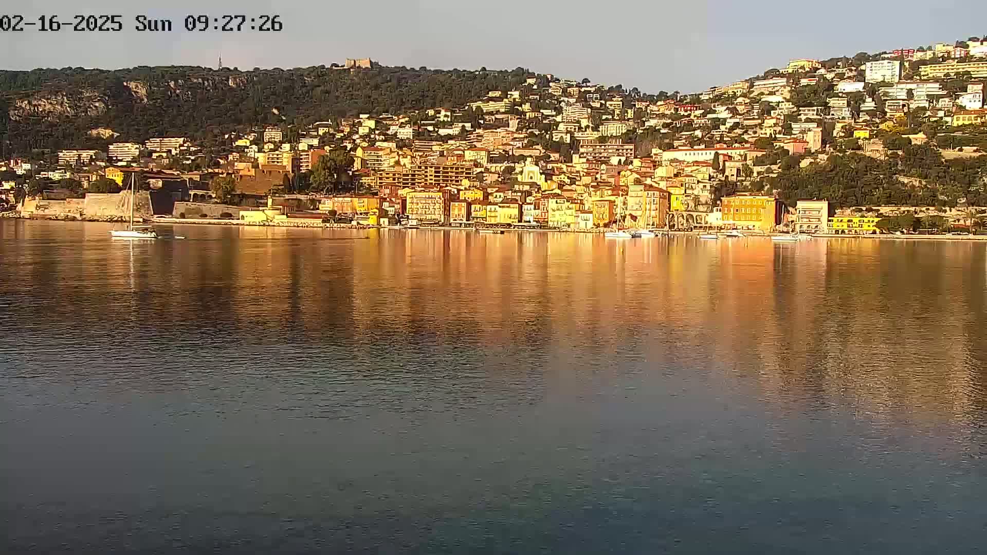 A calm body of water reflects a hillside town of colorful buildings under a clear sky.