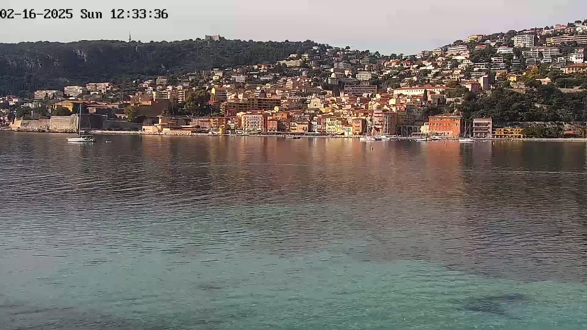 A picturesque coastal town, nestled against a hillside, is reflected in the calm waters of a bay under a clear sky.