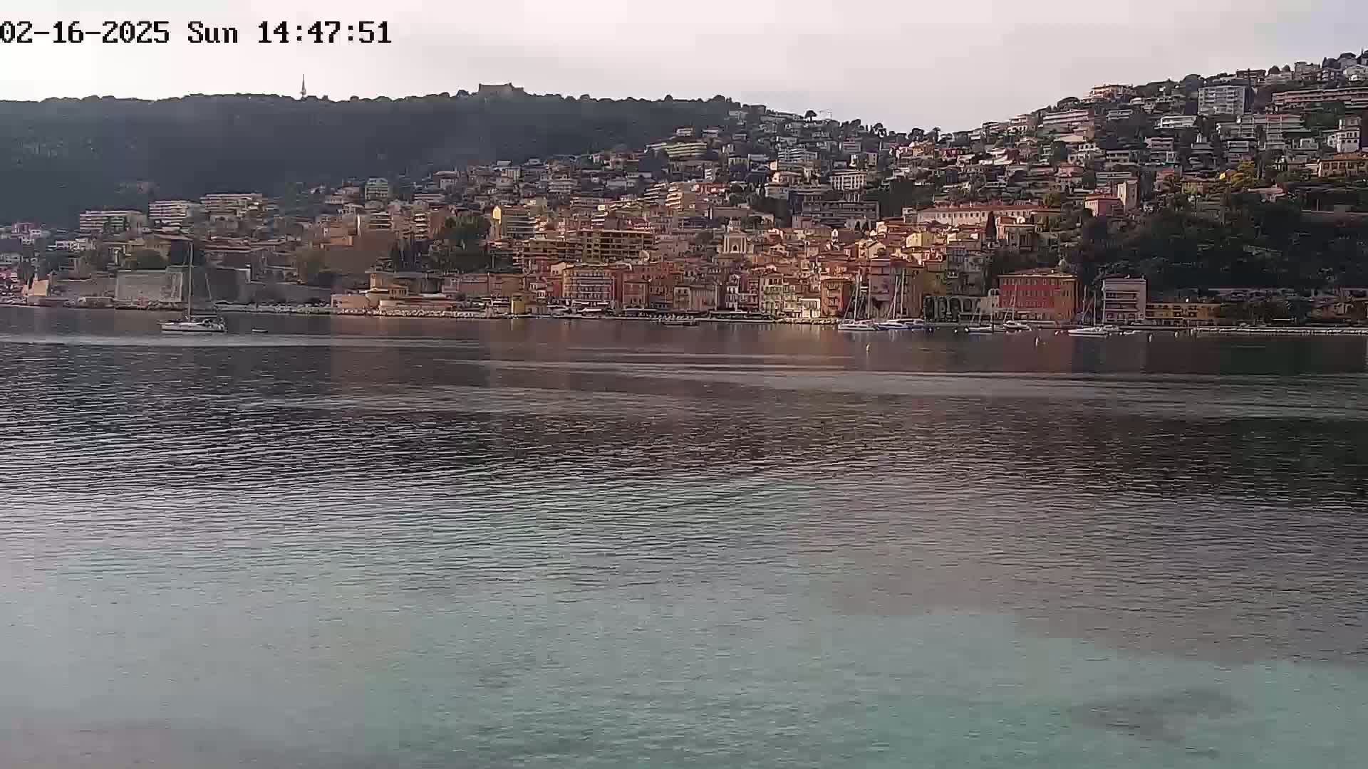 A calm body of water stretches out in front of a hillside town with numerous buildings, under a mostly clear sky.
