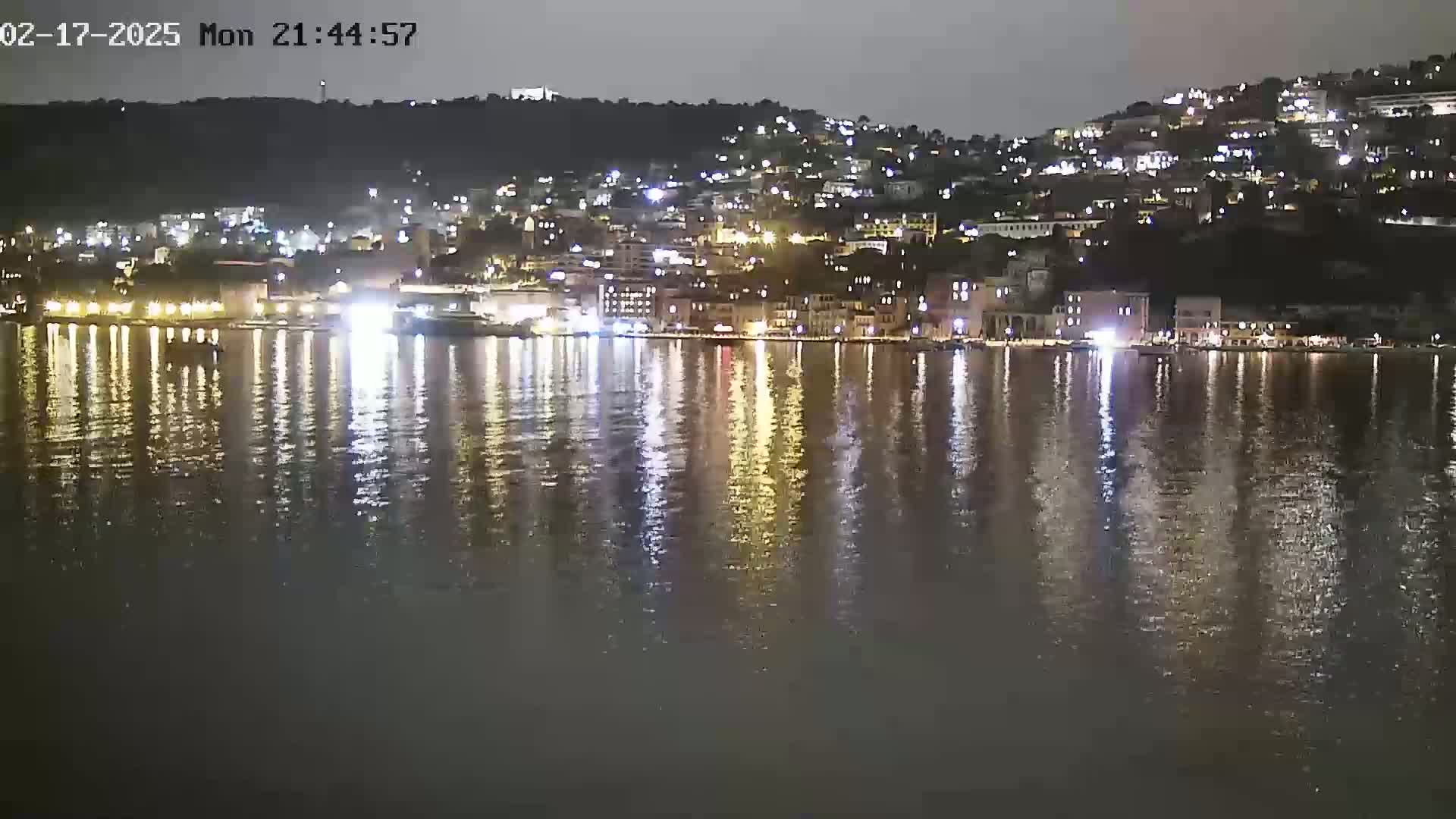A nighttime view of a city's waterfront, illuminated by lights reflecting on calm water.