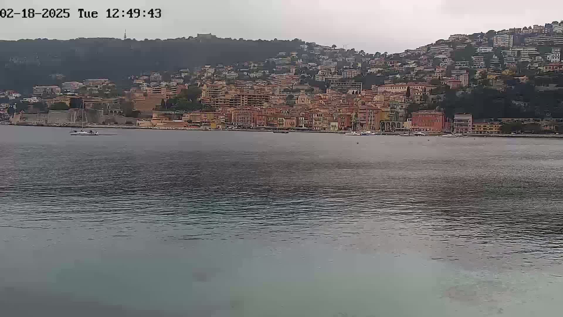 A coastal town with numerous buildings densely packed on a hillside is seen across a calm body of water under an overcast sky.