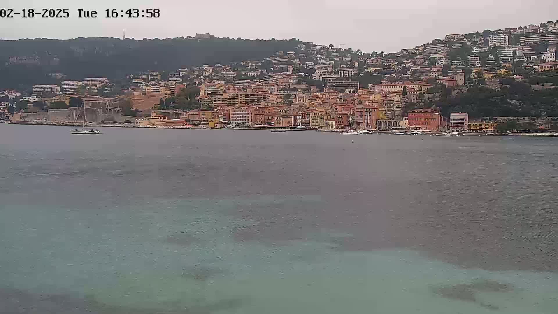 A coastal town with many buildings of varying heights is situated on a hillside overlooking a calm body of water under an overcast sky.
