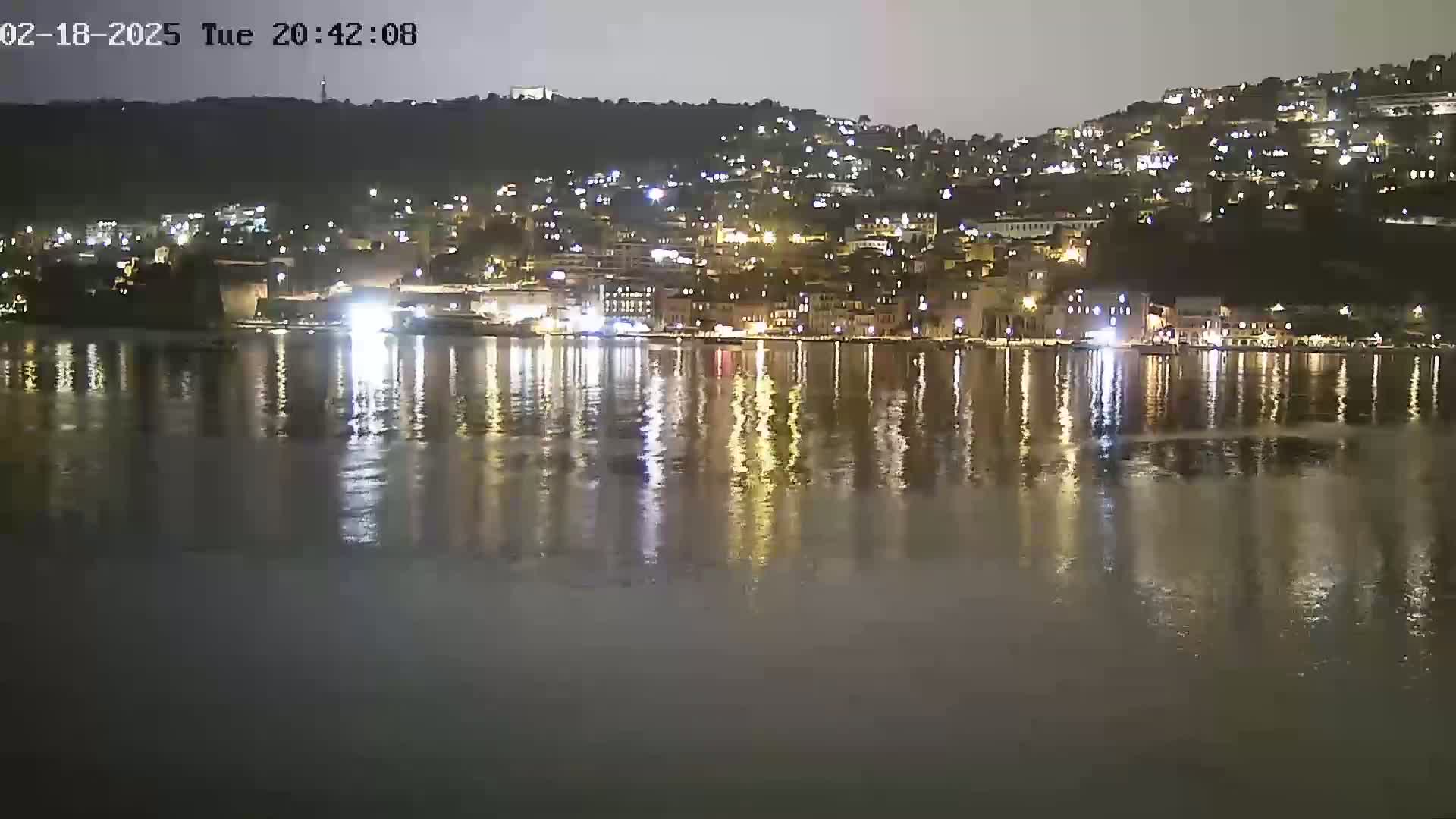 A nighttime view of a city on a hill overlooking calm water, reflecting the lights of the buildings.