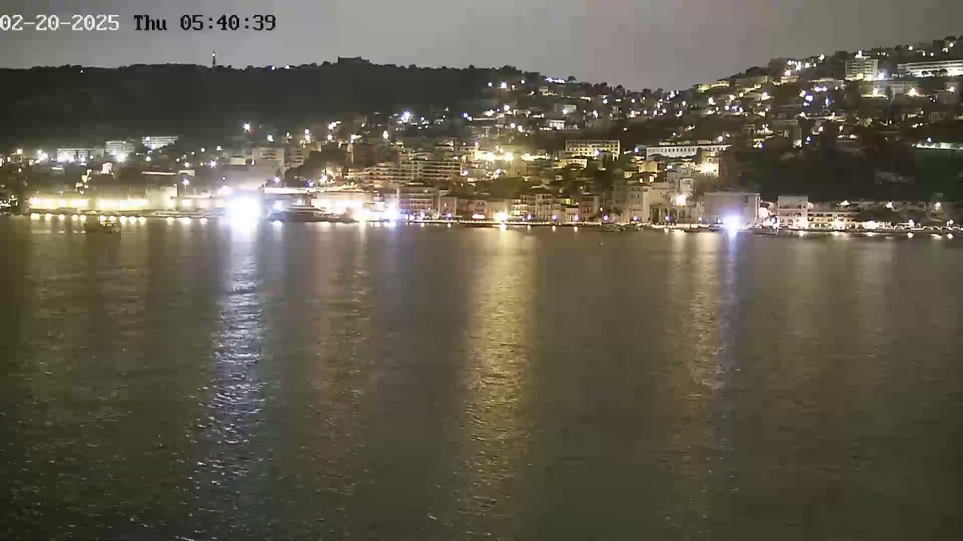 A nighttime view of a city's waterfront, with its buildings illuminated, reflected in calm water.