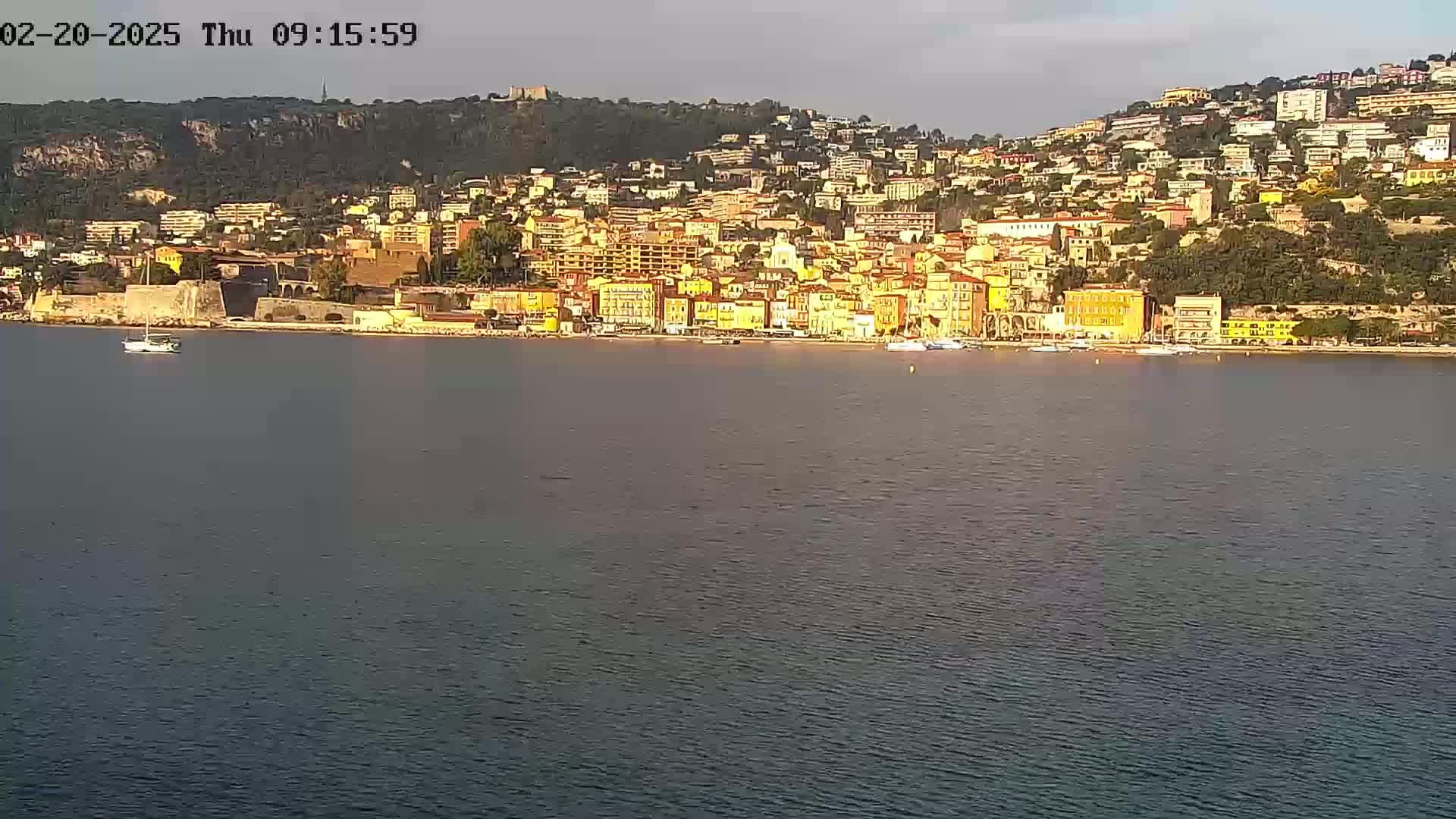 A coastal town with yellow buildings sprawls across a hillside, overlooking a calm body of water under a mostly clear sky.
