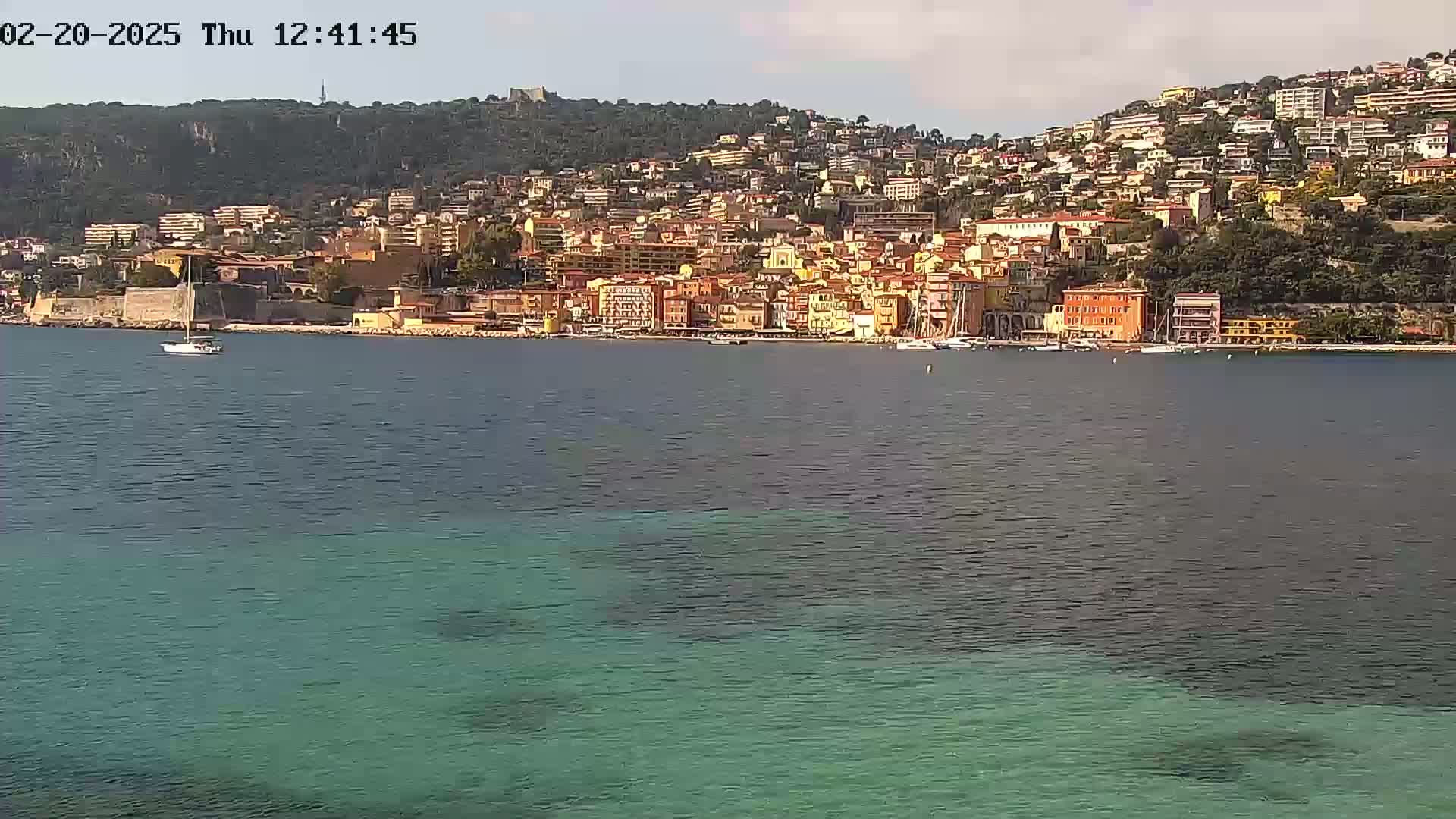 A sailboat is visible in the calm water in front of a coastal town built on a hillside under a mostly clear sky.