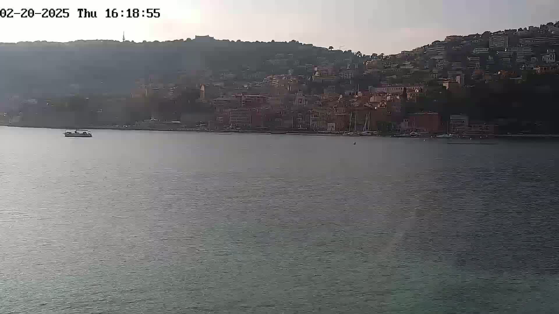 A small boat is on calm water in front of a hillside town under a mostly clear sky.
