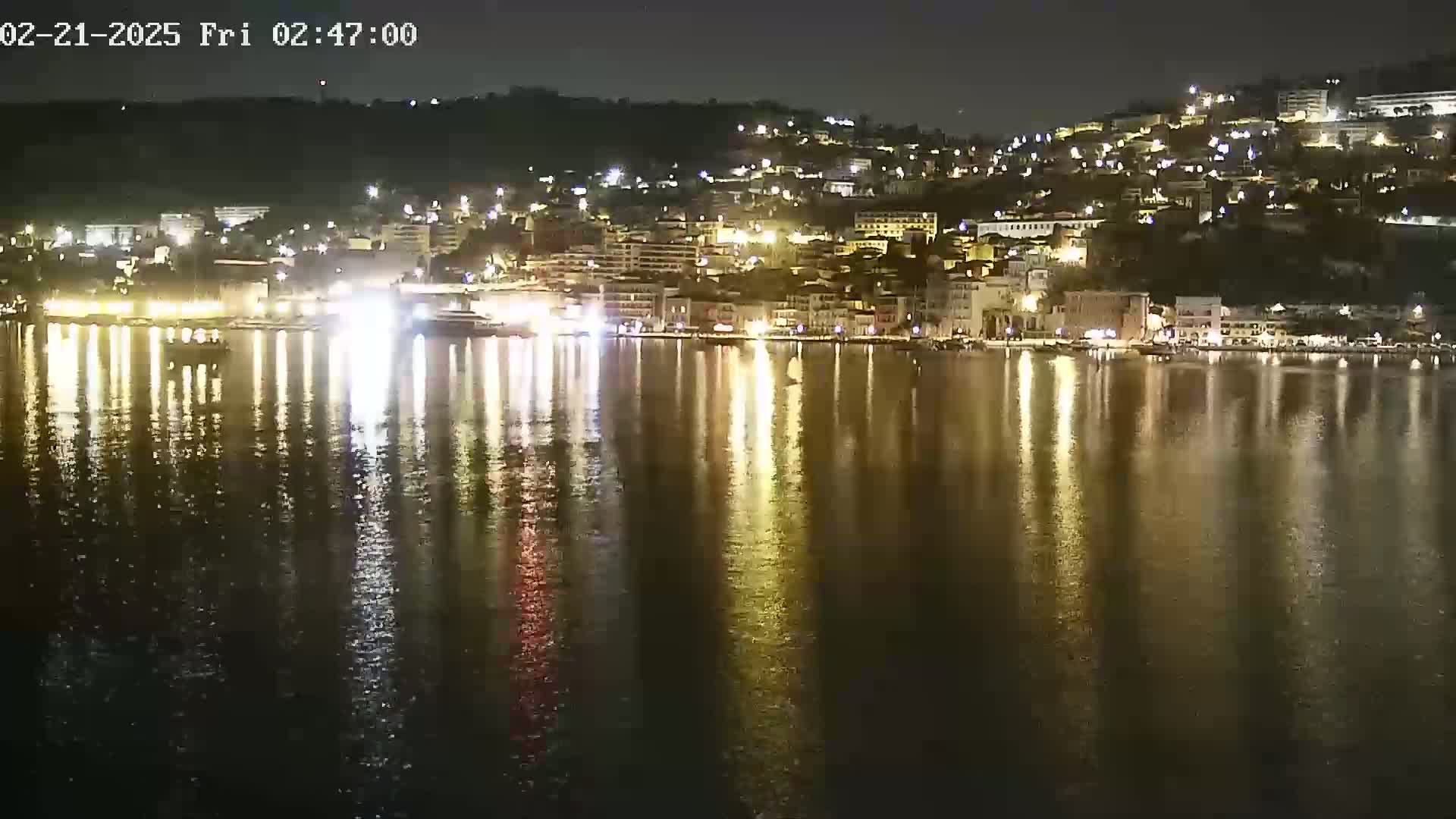 A nighttime view of a city's waterfront, illuminated by numerous lights reflecting on the calm water.