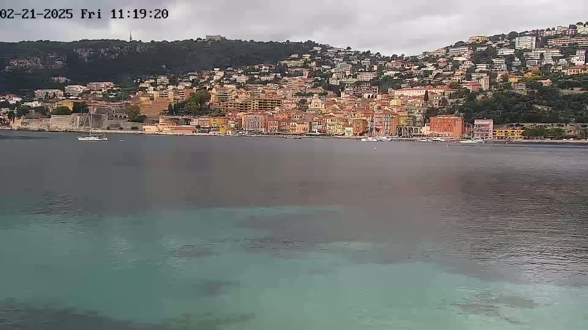 A sailboat is visible in the calm waters of a bay, with a hillside town of colorful buildings under a cloudy sky.