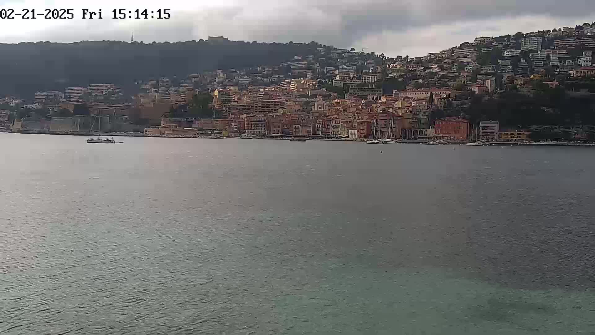 A boat is on the water in front of a hillside town under an overcast sky.