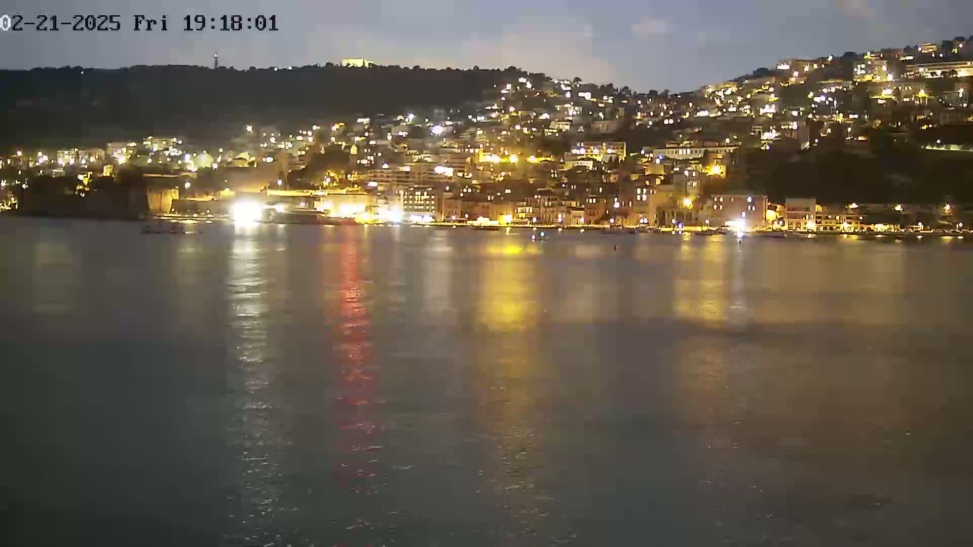 A nighttime view of a city on a hill overlooking a calm body of water, with lights illuminating the buildings and their reflections visible on the water's surface.