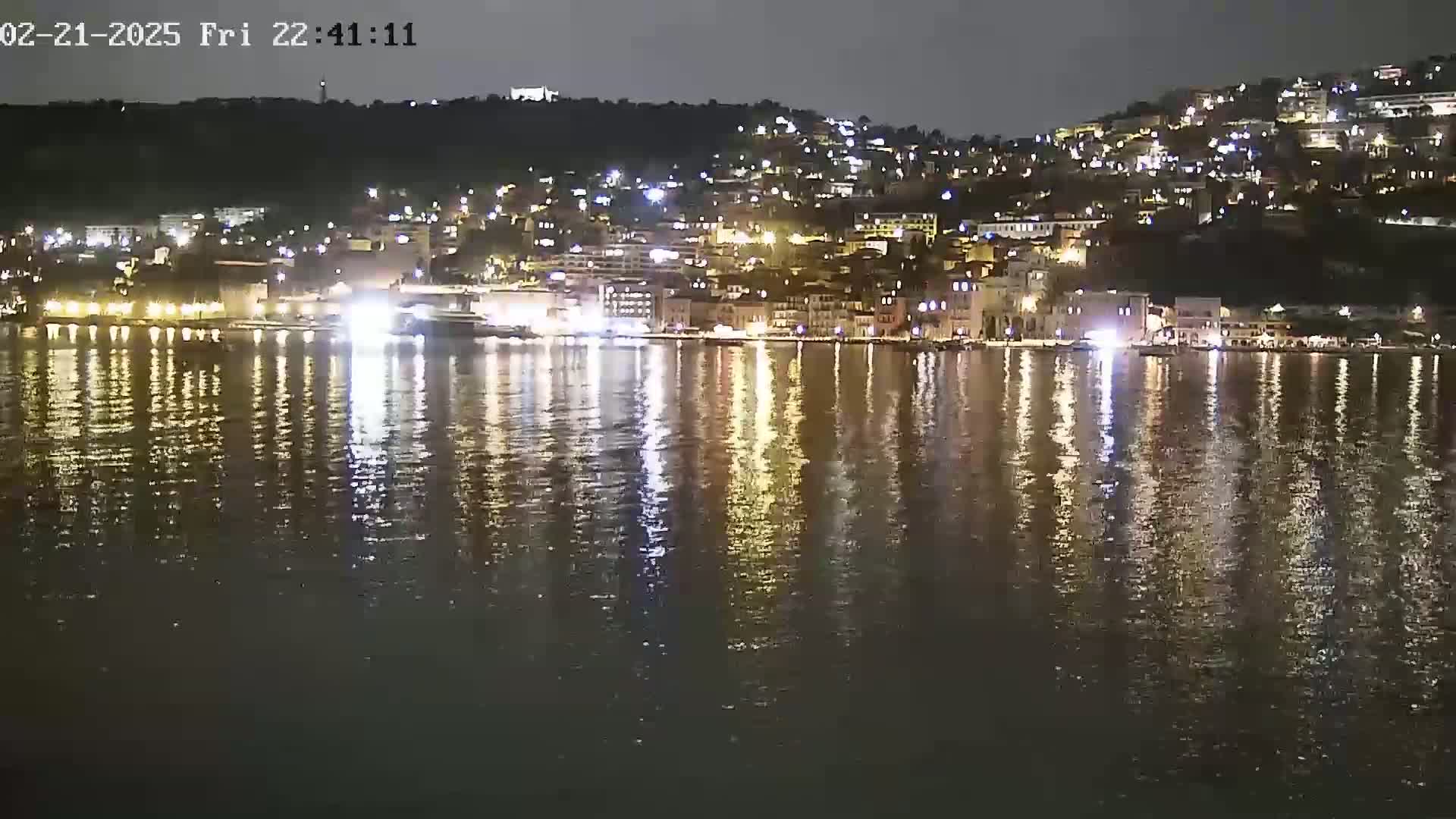 A nighttime view of a town's waterfront, illuminated by many lights, reflecting on a calm body of water.