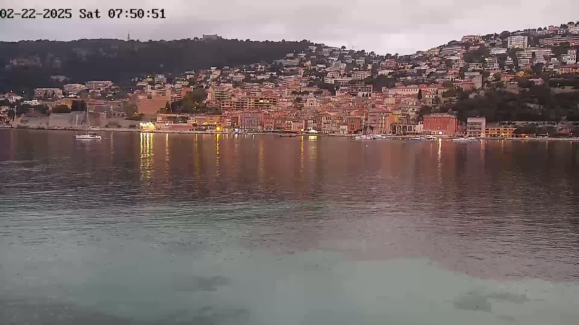 A town of closely clustered buildings on a hillside is reflected in the calm water of a harbor at twilight.