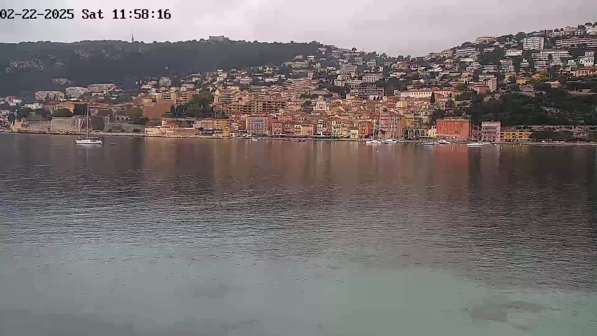 A calm body of water reflects a hillside town of closely clustered buildings under an overcast sky.