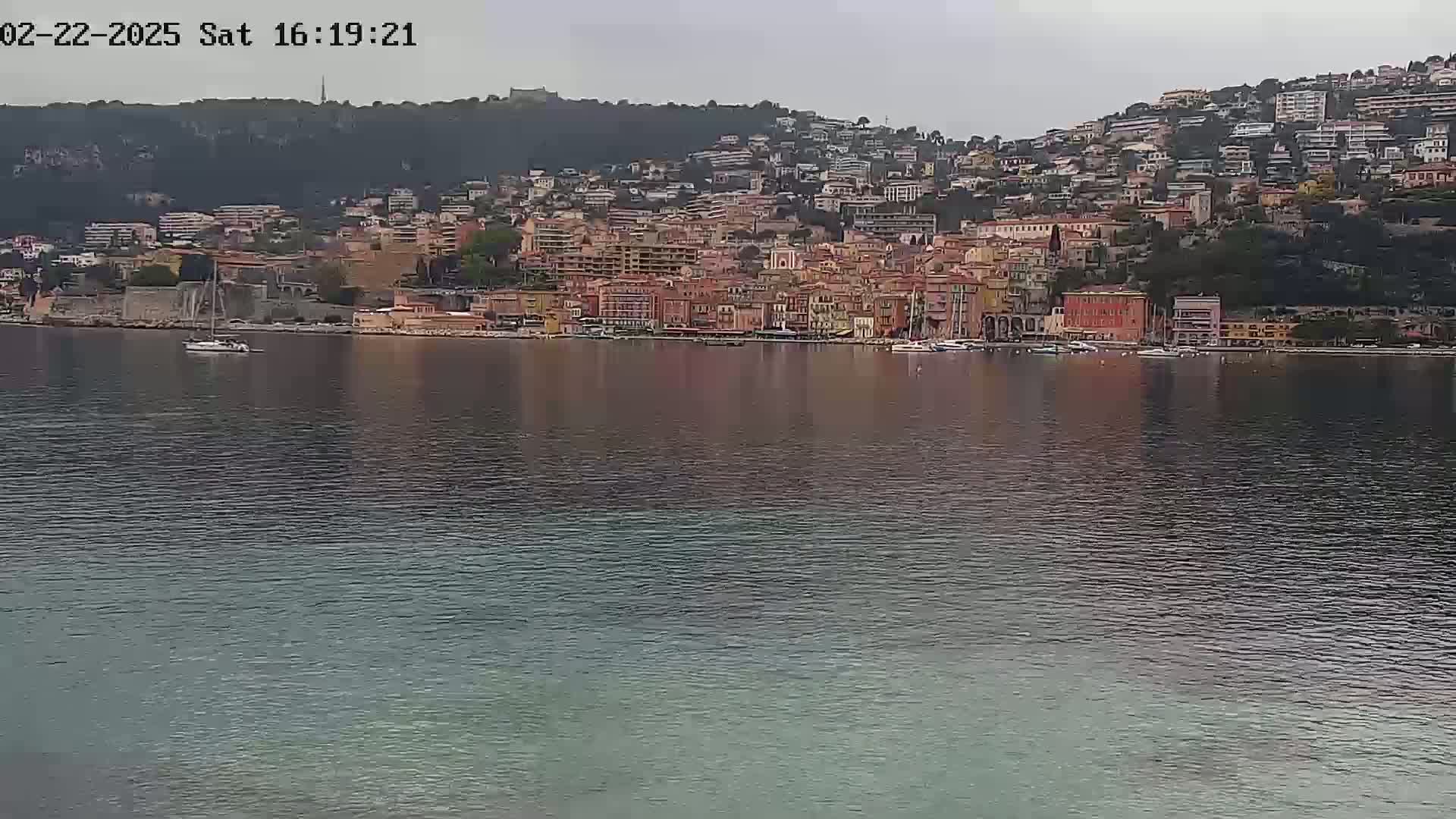 A waterfront town with many closely clustered buildings on a hillside is reflected in the calm, gray water under an overcast sky.