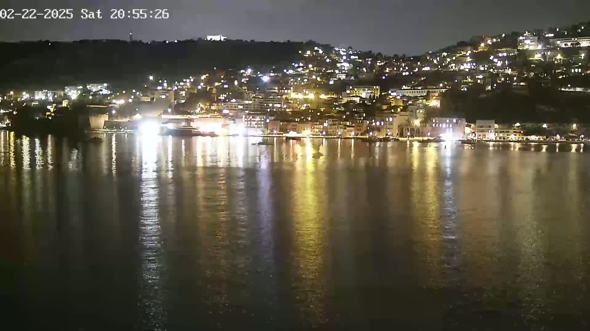 A nighttime view of a town's waterfront, illuminated with lights, reflected on calm water.