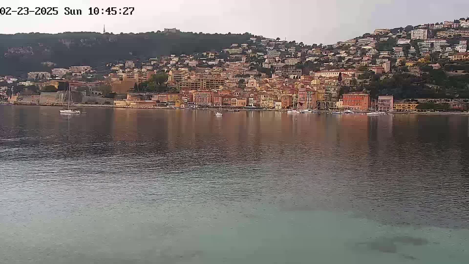 A calm body of water reflects a hillside town of closely packed buildings under an overcast sky.