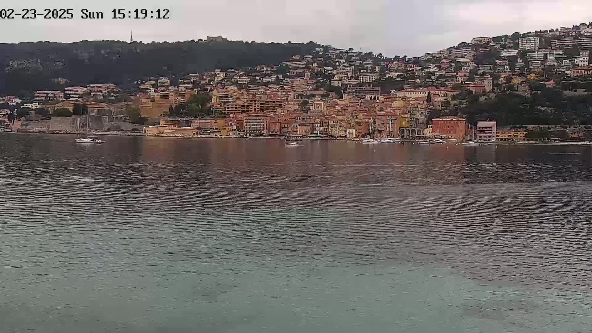 A hillside town of tan and orange buildings sits on a calm body of water under an overcast sky.