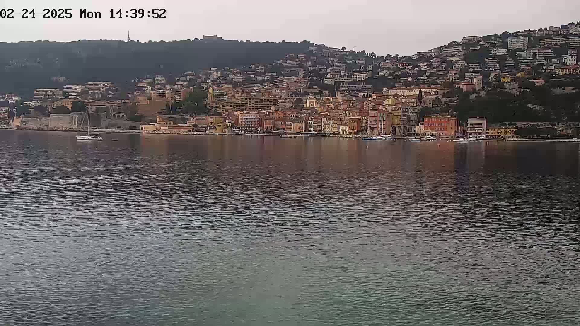 A coastal town with numerous buildings of varying heights and colors is situated on a hillside overlooking a calm body of water under an overcast sky.