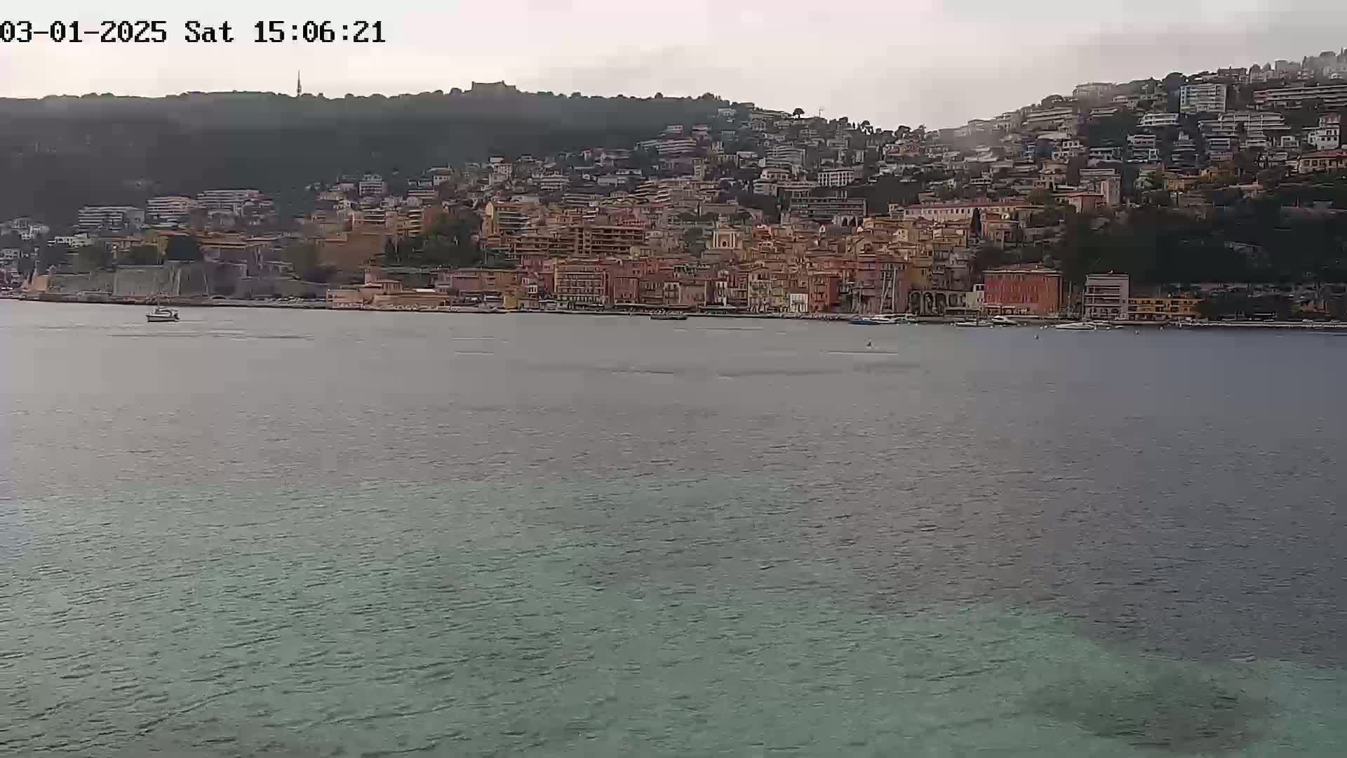 A coastal town with many buildings on a hillside is seen across a calm body of water under an overcast sky.