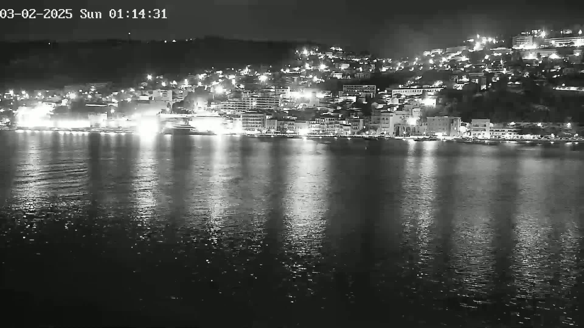 A nighttime, low-angle view shows a town's brightly lit waterfront along a calm body of water.