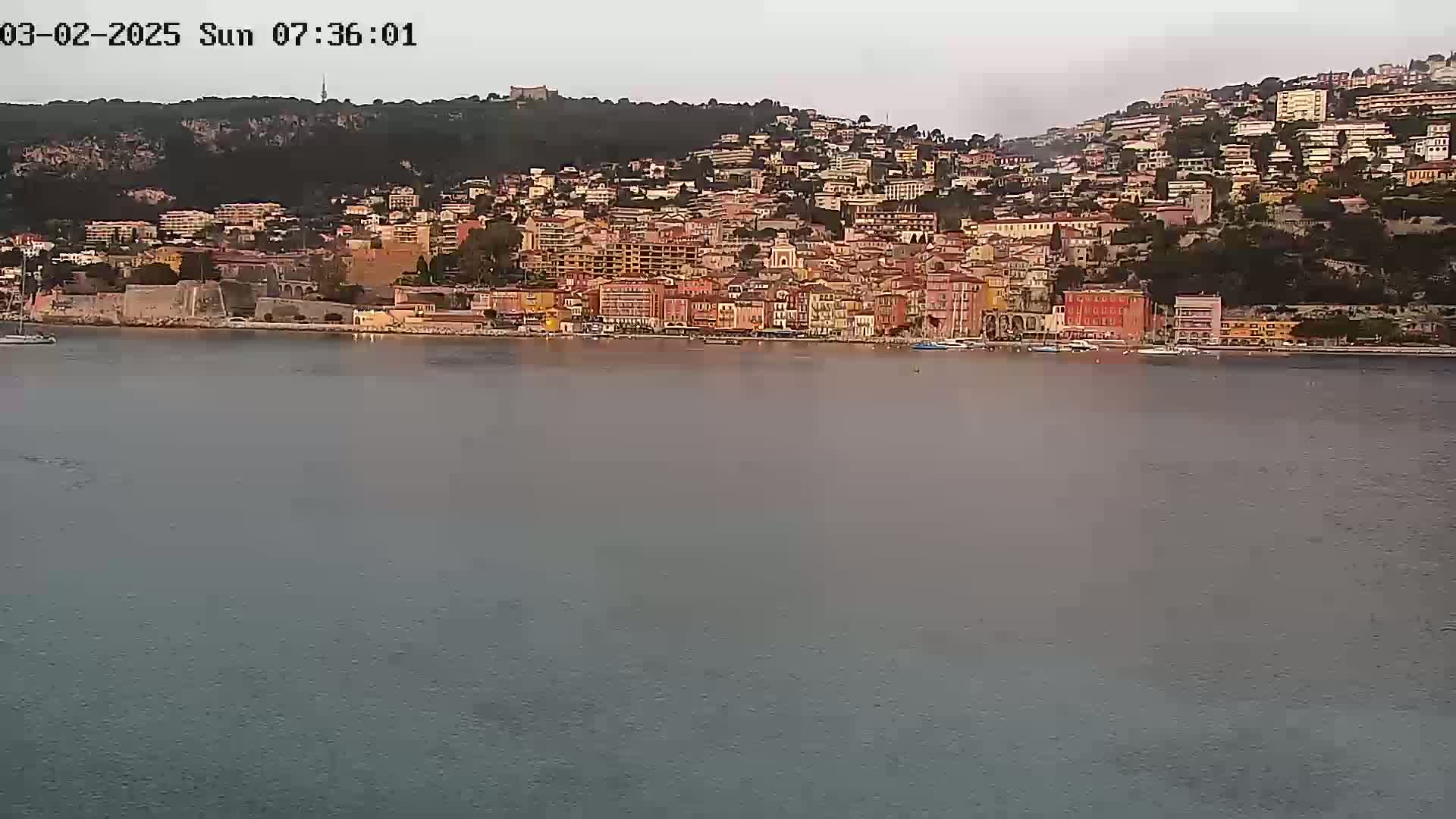 A coastal town with numerous buildings of varying heights and colors sits on a hillside overlooking a calm body of water under a hazy sky.