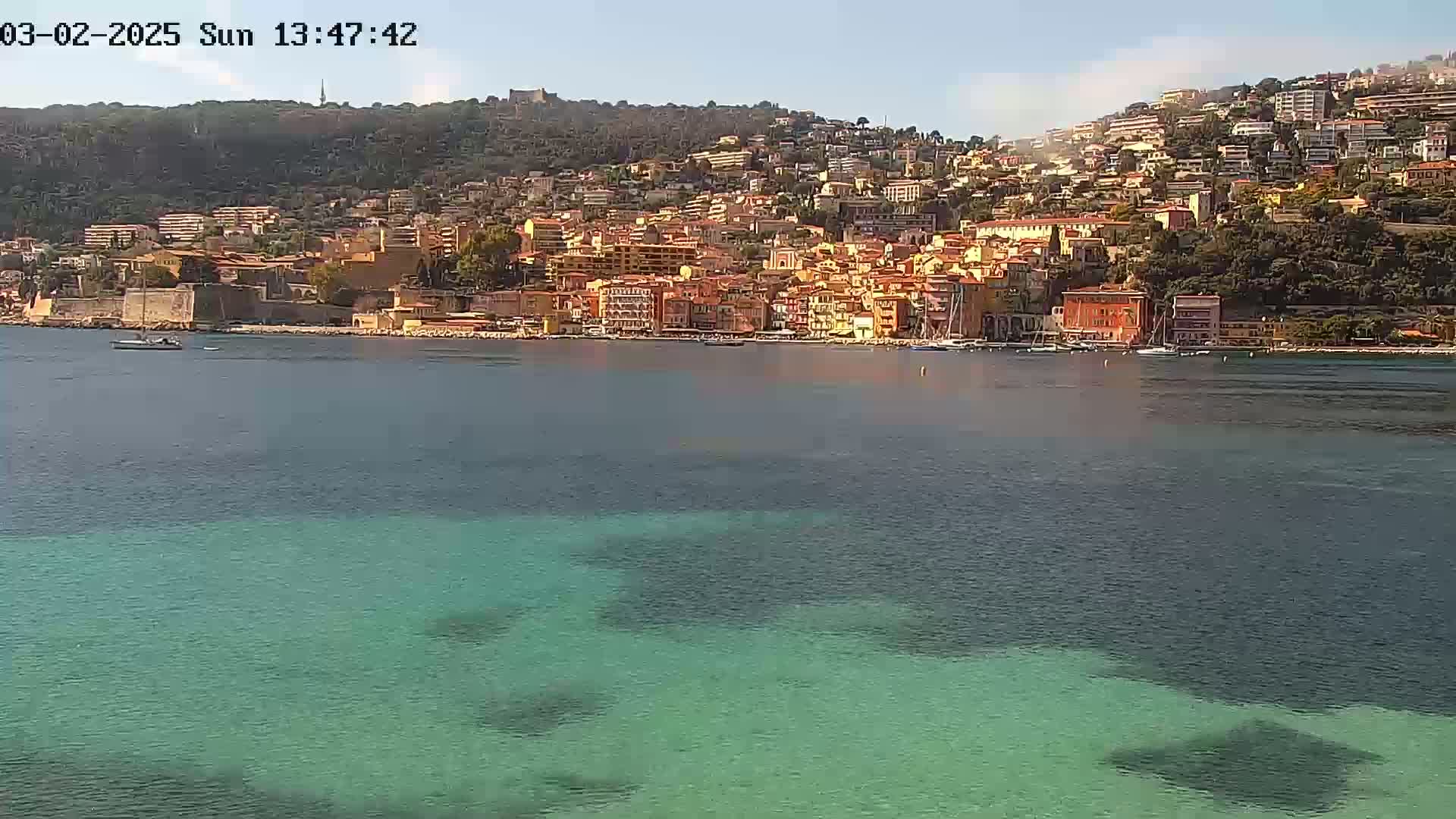 A coastal town with colorful buildings sits on a hillside overlooking a calm, clear teal sea on a sunny day.