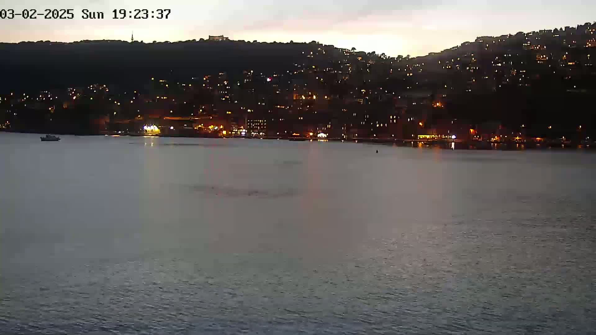 A nighttime view of a calm body of water with a hill covered in illuminated buildings in the background under a dusky sky.