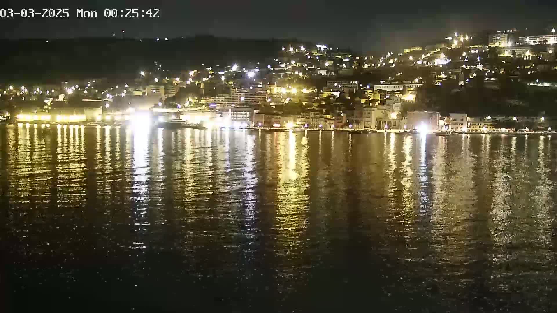 A nighttime view of a waterfront town with many lights reflected in calm water.