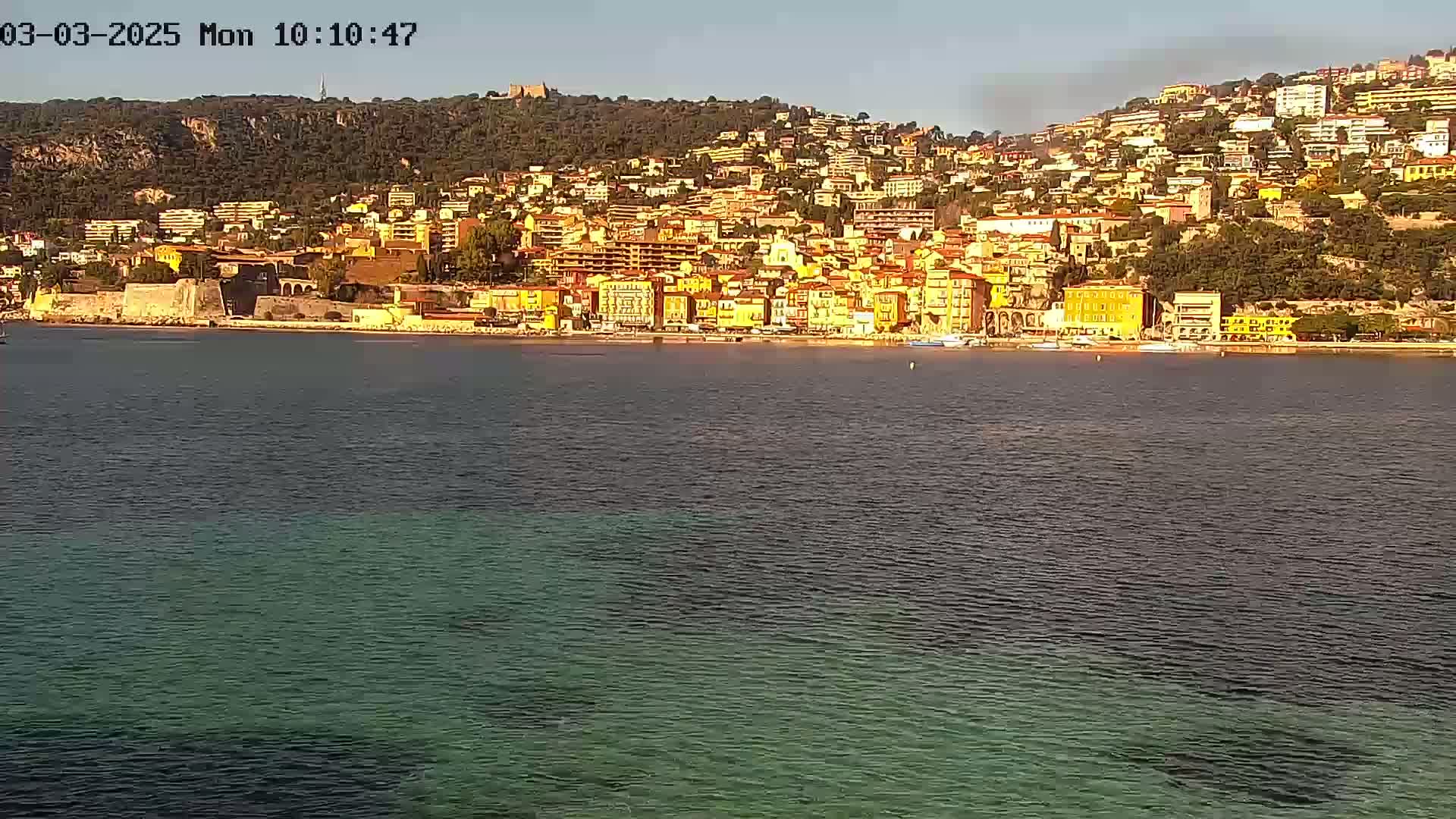 A coastal town with numerous buildings on a hillside, overlooking calm, clear water under a sunny sky.