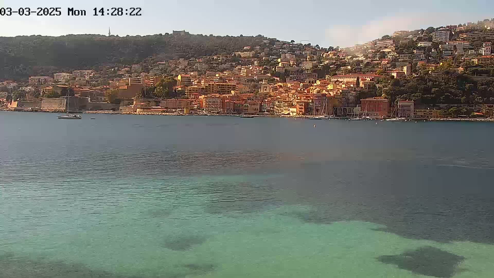 A coastal town with numerous buildings of varying heights and colors is situated on a hillside overlooking a calm body of water with clear, shallows near the shore.