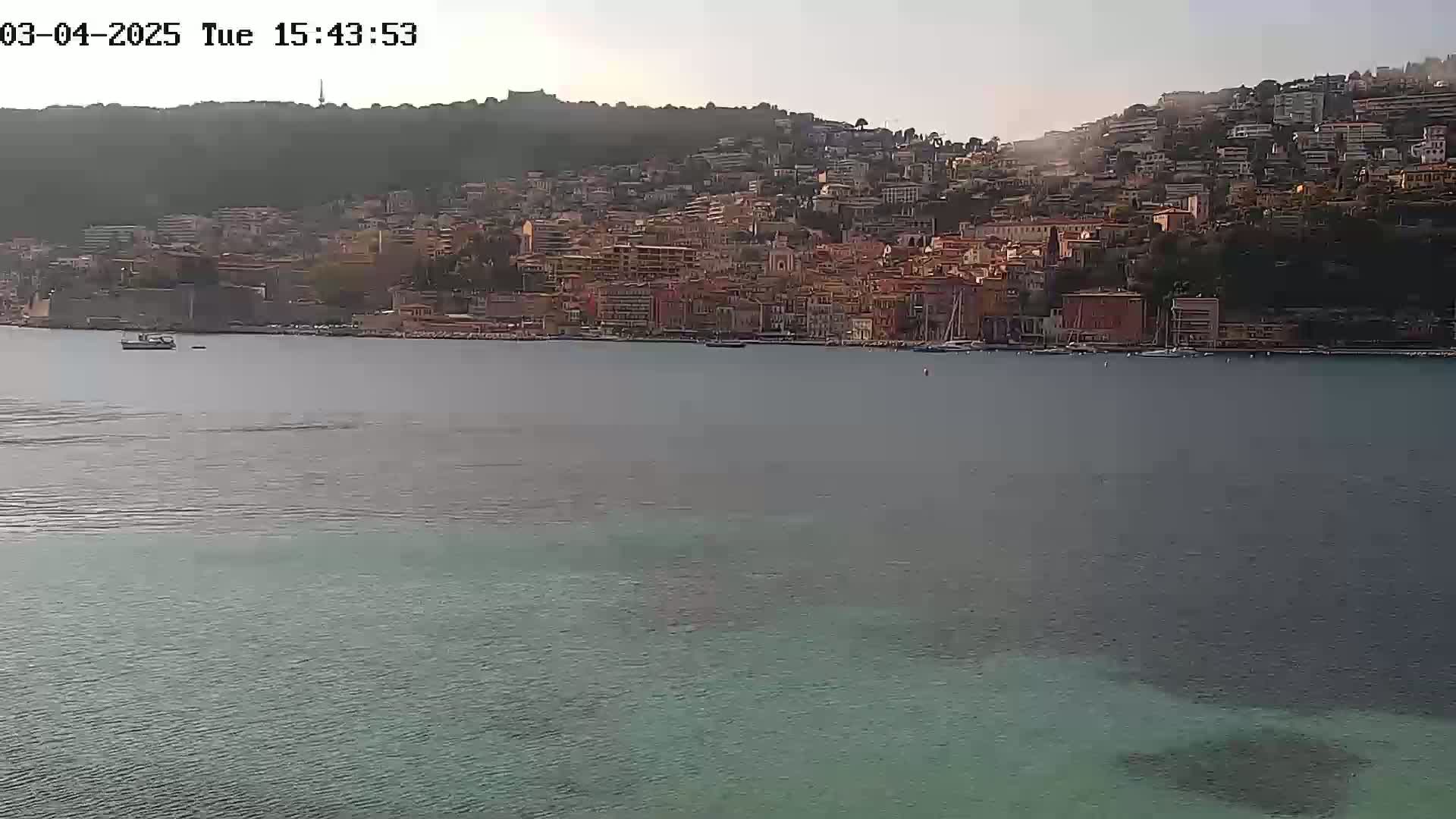 A coastal town built on a hillside is visible across a calm body of water under a hazy sky.