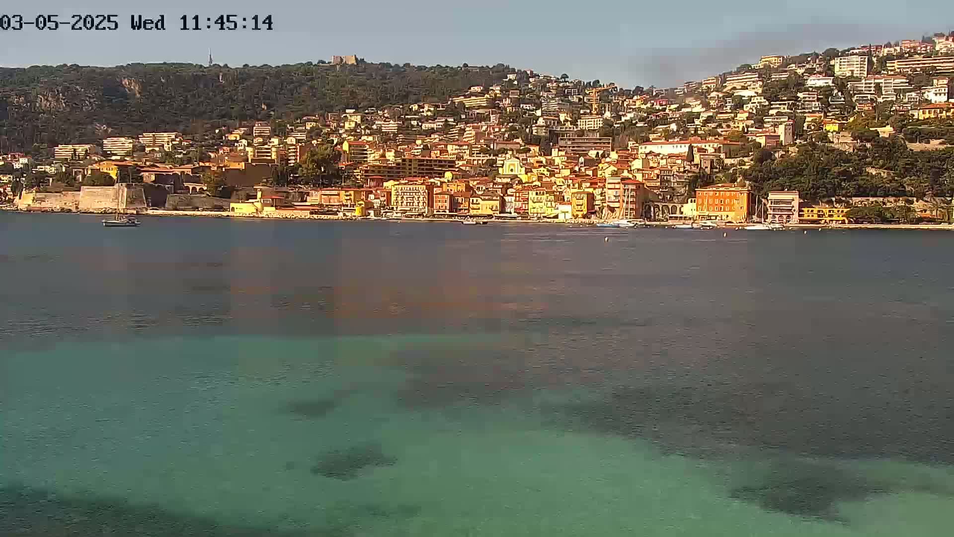 A coastal town with numerous buildings of varying heights and colors sits on a hillside overlooking a calm body of teal water under a clear sky.